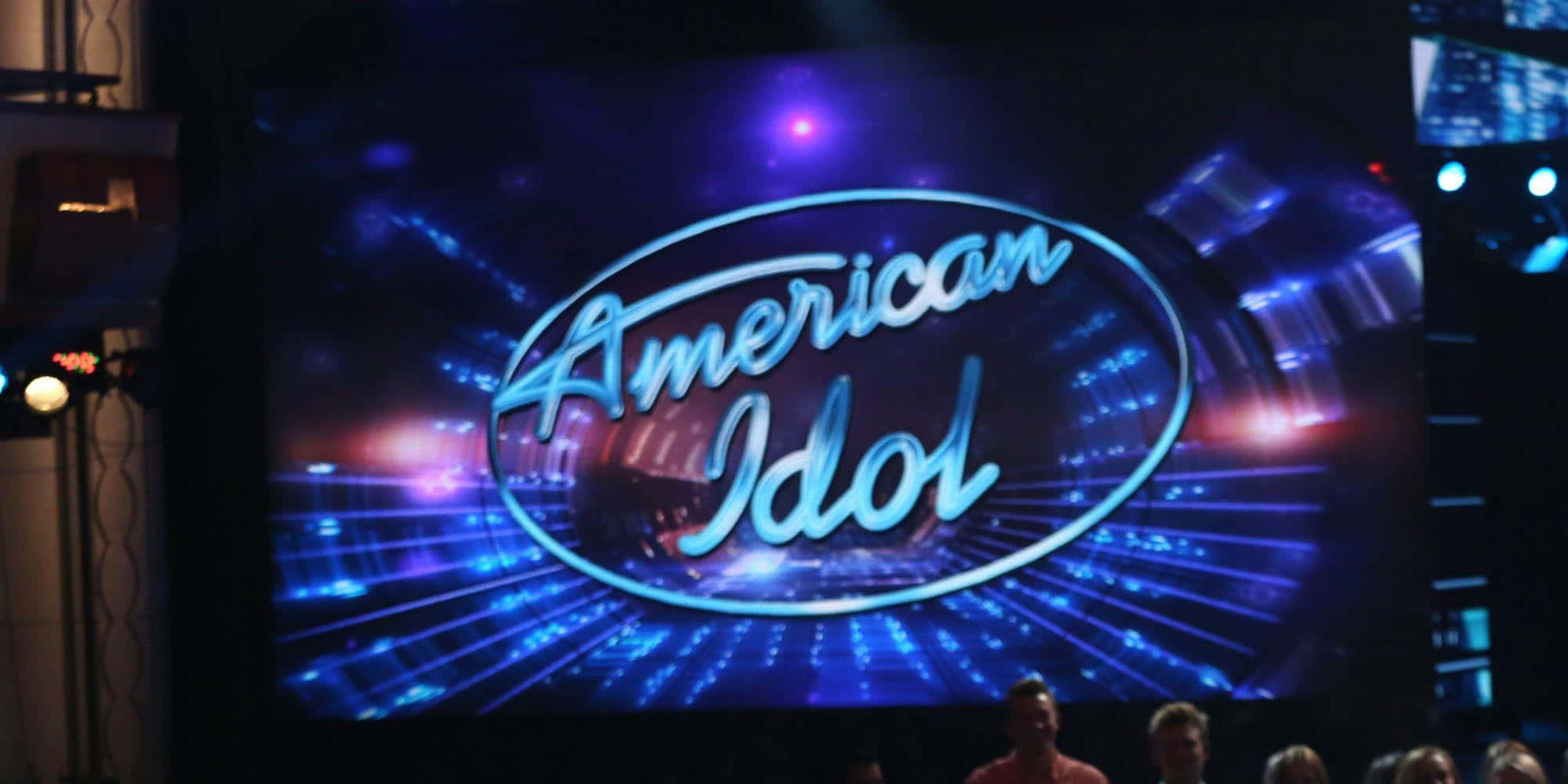 american idol 桌布 壁紙 — 大螢幕顯示發光的American Idol標誌，藍色和紫色的舞台燈光向外輻射。