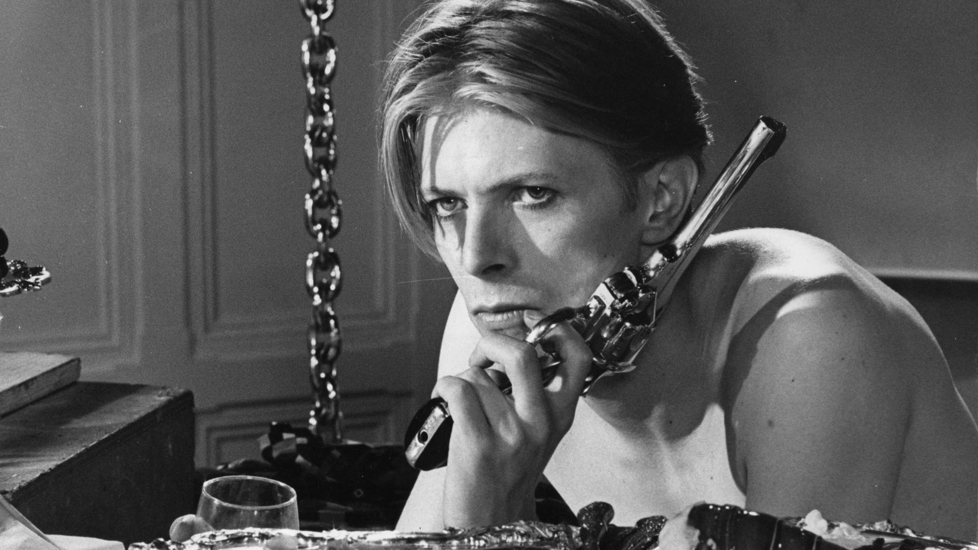 David Bowie 桌布 壁紙 — 大衛·鮑伊在黑白鮮明的場景中，手持左輪手槍抵住下巴，眼神專注。