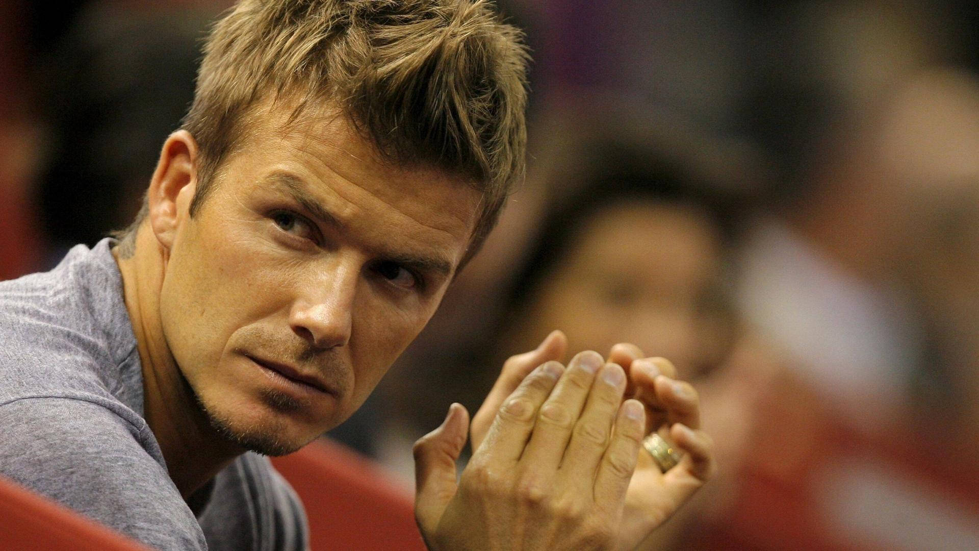 David Beckham 桌布 壁紙 — 大衛貝克漢在模糊的球場背景下，雙手合十，戴著金戒指，凝視著。