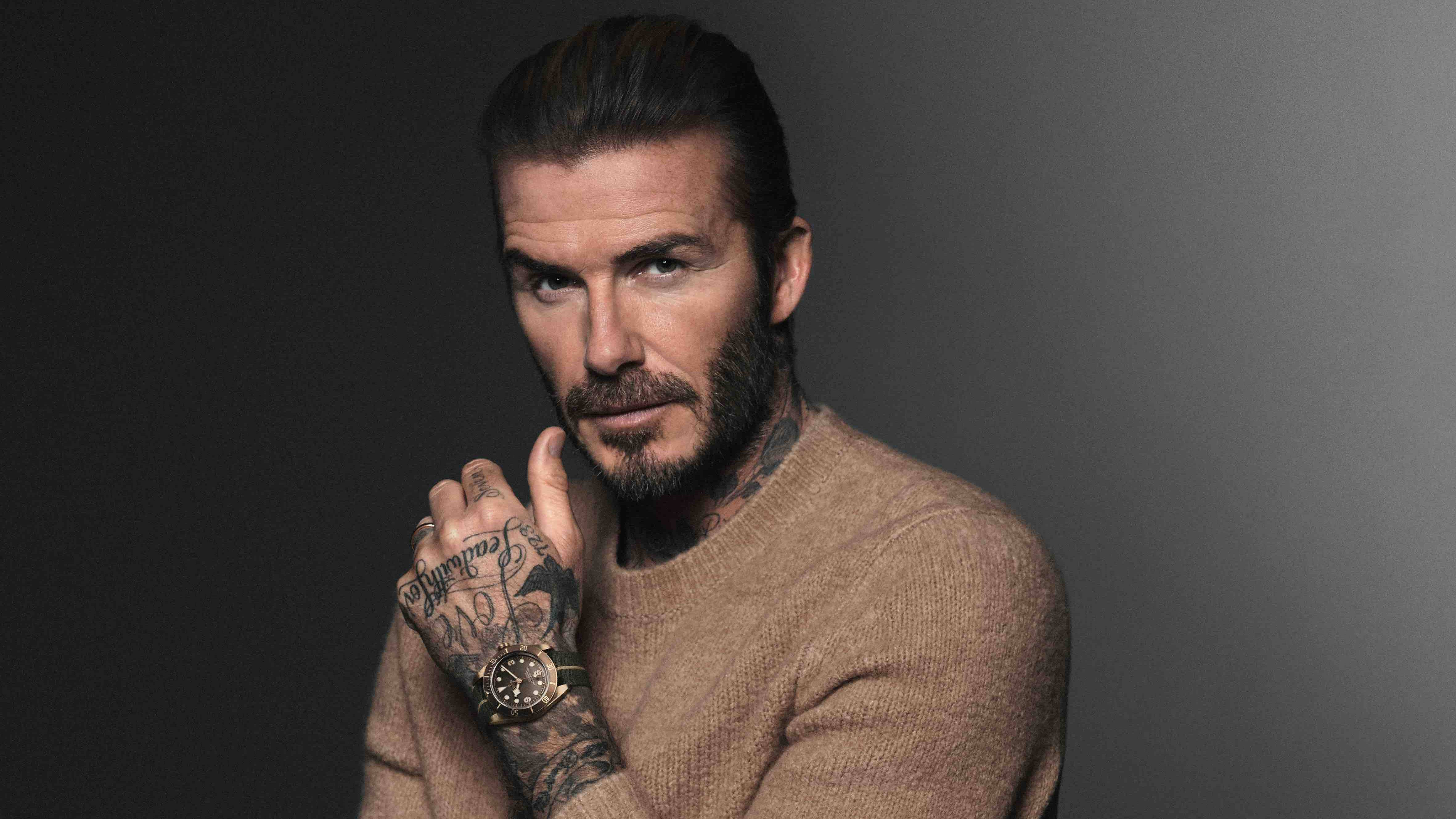 David Beckham 桌布 壁紙 — 大衛貝克漢在深灰色工作室背景前自信地擺姿勢，身穿米色毛衣，露出刺青和手錶。
