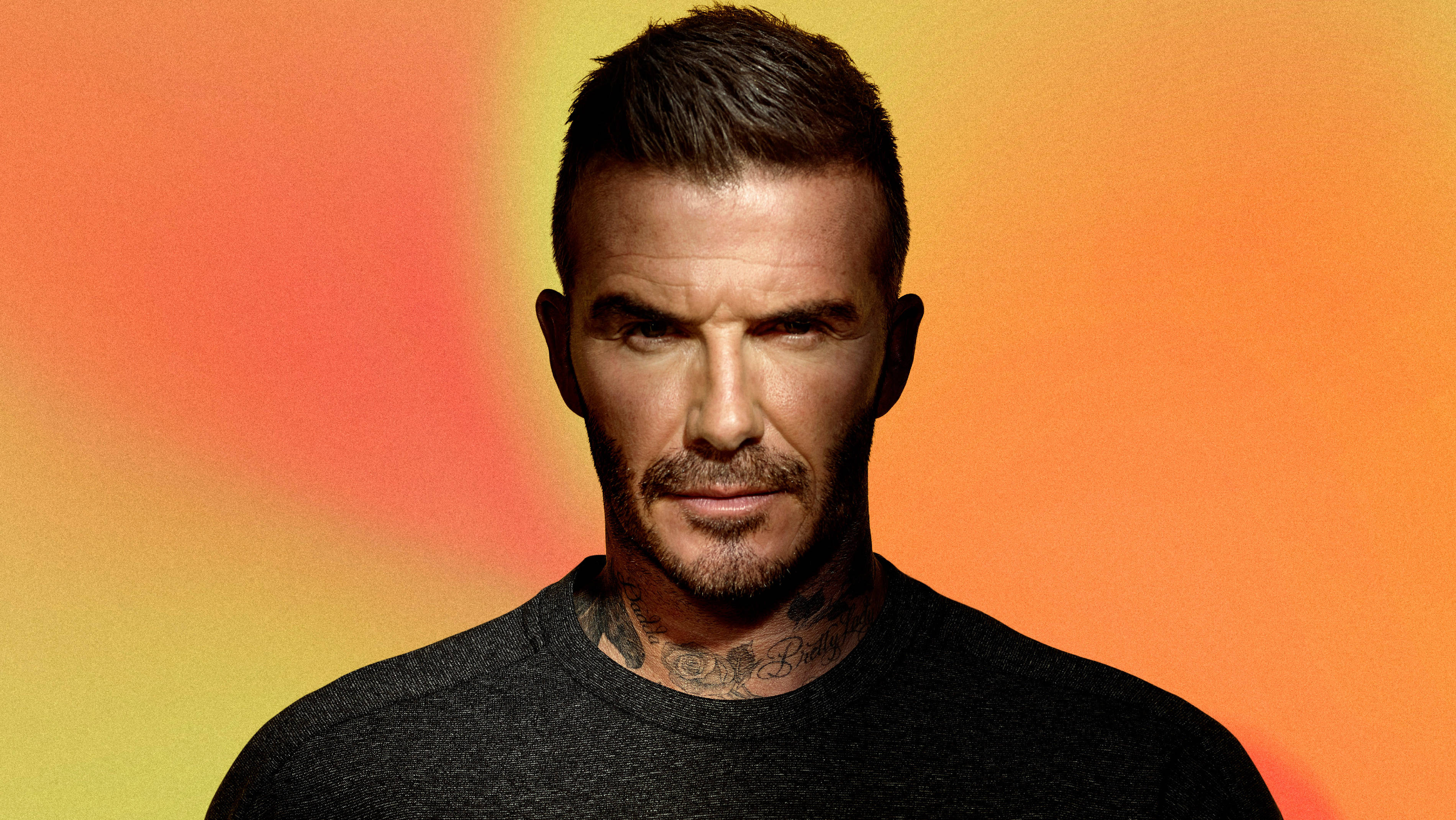 David Beckham 桌布 壁紙 — 大衛貝克漢在溫暖的橙黃漸變背景前凝視，頸部紋身清晰可見。