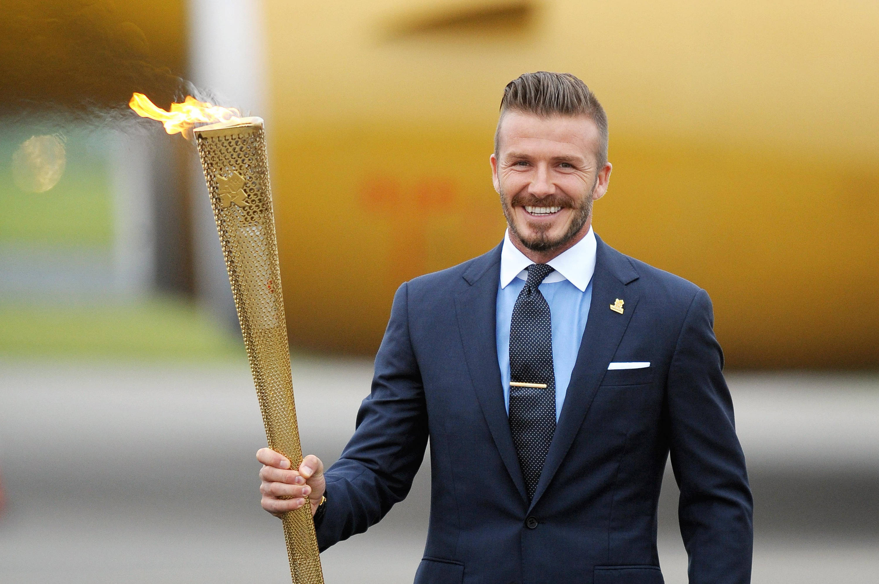 David Beckham 桌布 壁紙 — 大衛貝克漢自豪地微笑，手持金色奧林匹克火炬，火焰明亮，背景為模糊的黃色。