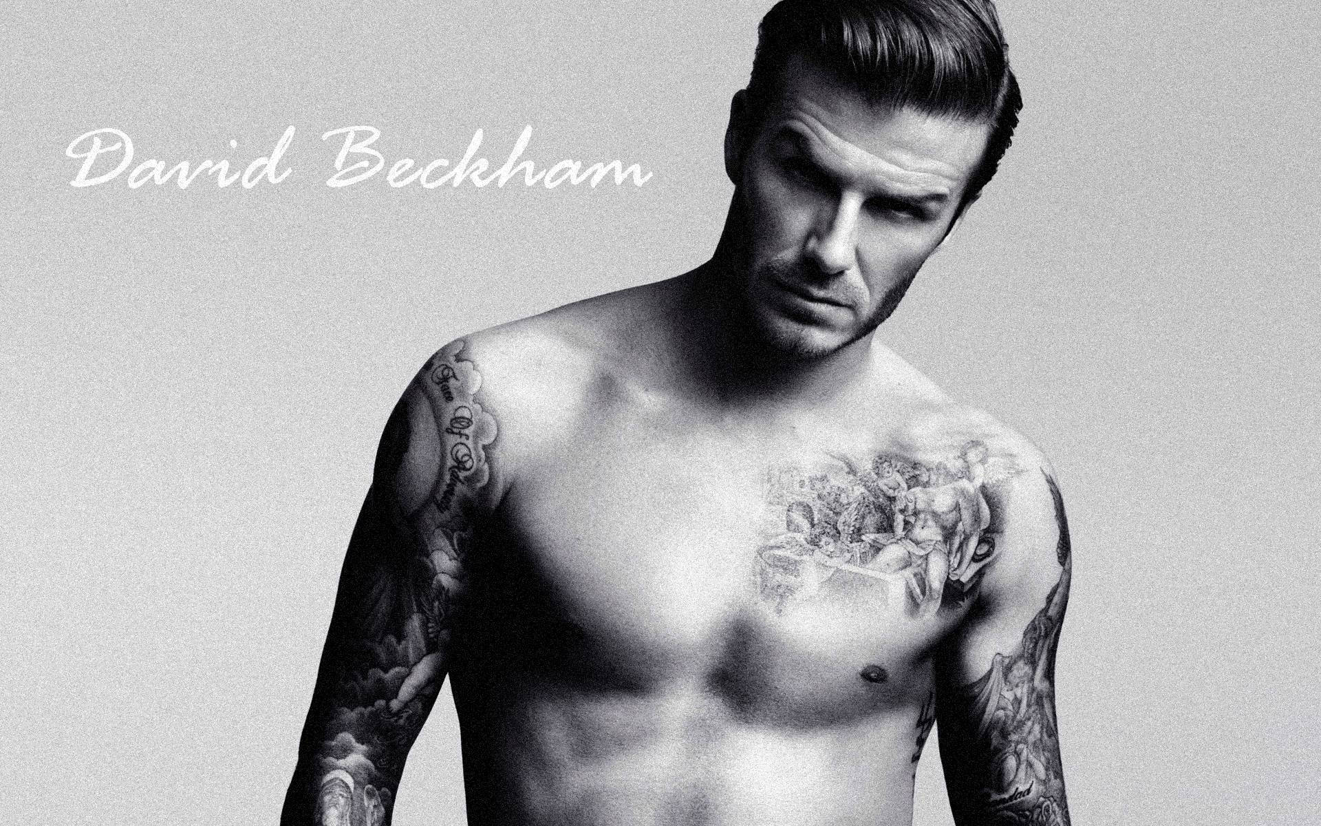 David Beckham 桌布 壁紙 — 大衛貝克漢赤裸上身，展示覆蓋手臂和胸部的複雜刺青，背景為純灰色。