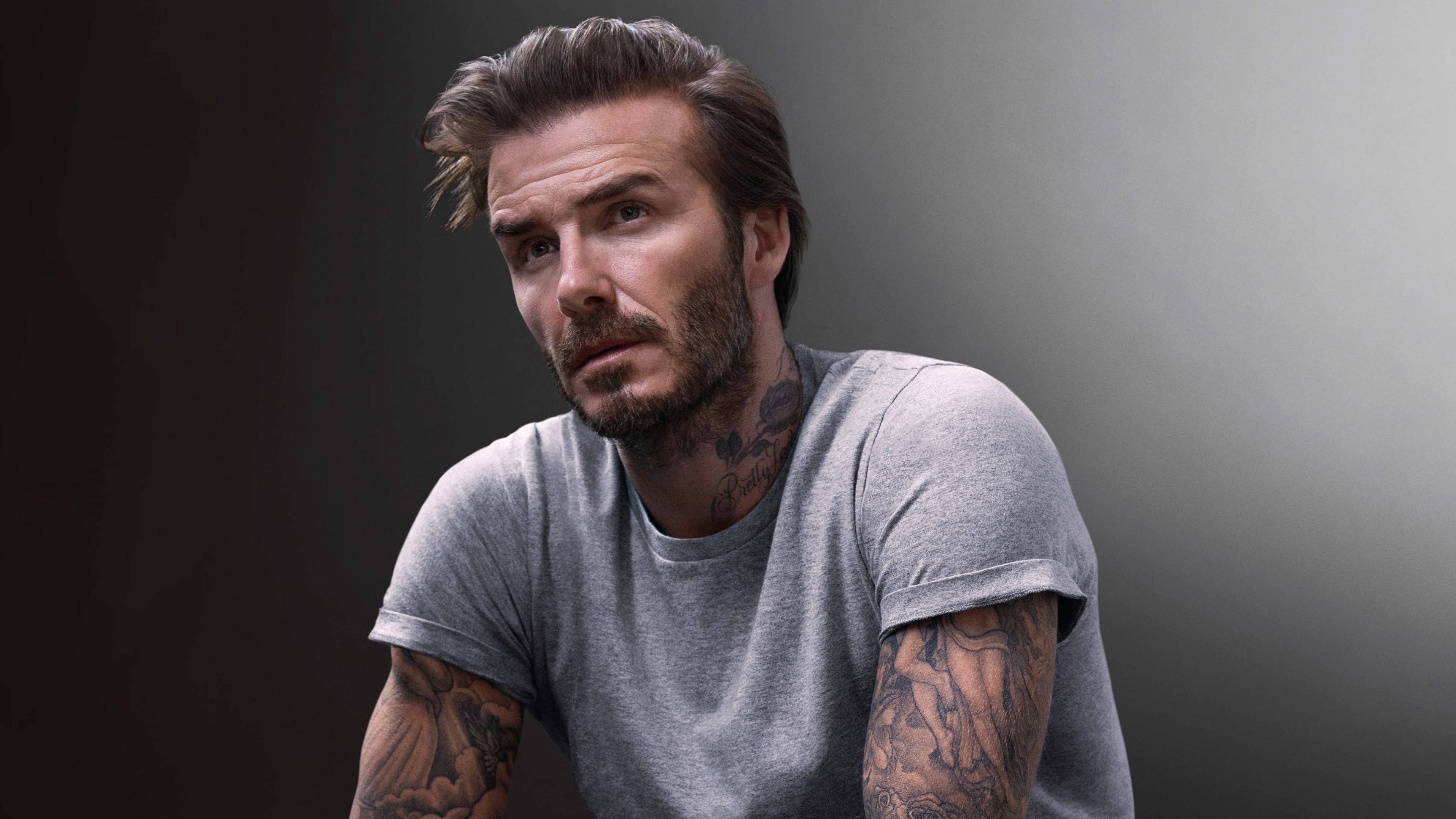 David Beckham 桌布 壁紙 — 大衛貝克漢身穿灰色T恤，手臂和頸部有刺青，背景為深色漸變。