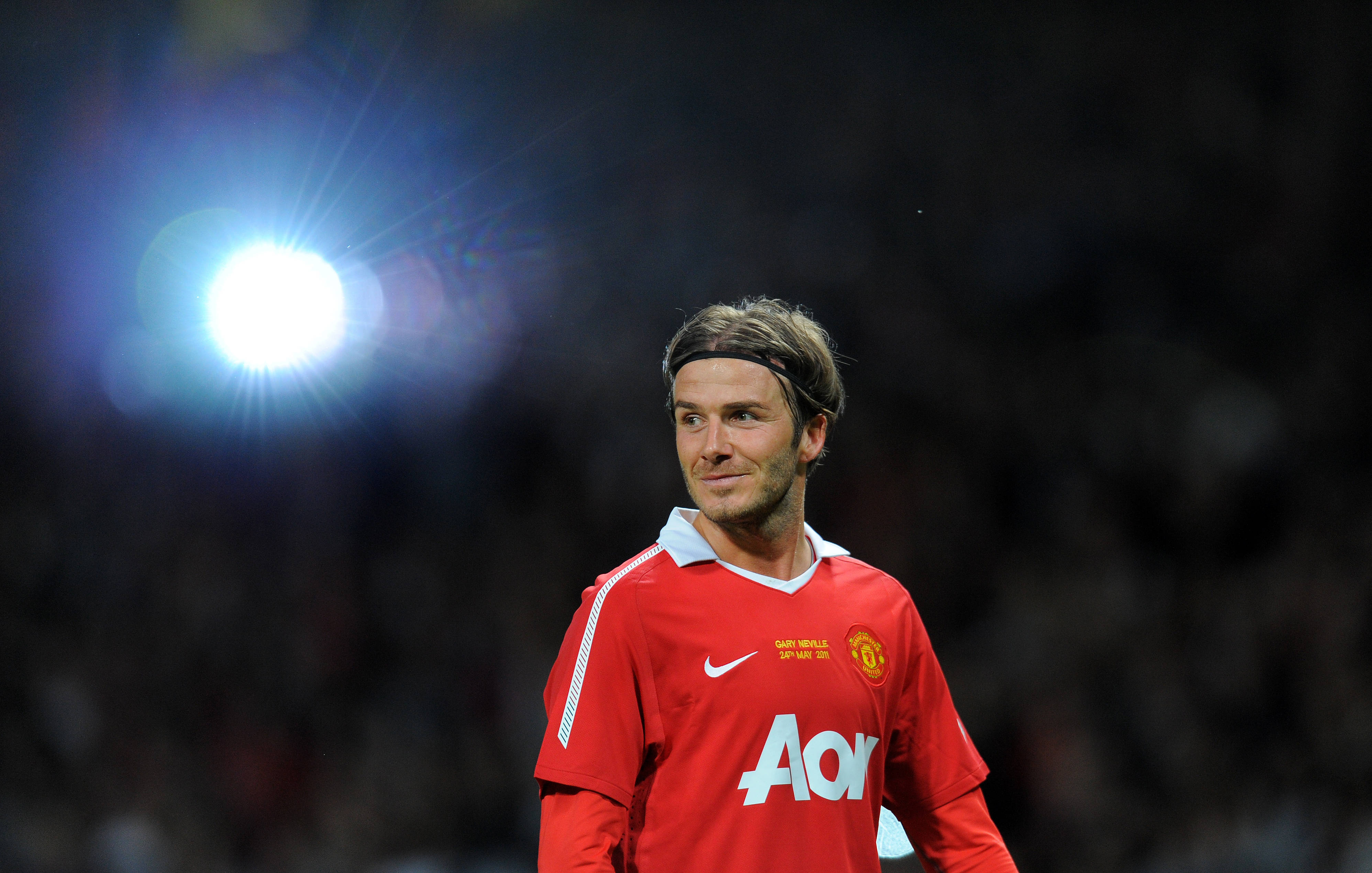 David Beckham 桌布 壁紙 — 大衛貝克漢身穿紅色曼聯球衣，站在明亮的球場燈光下，背後是模糊的人群。