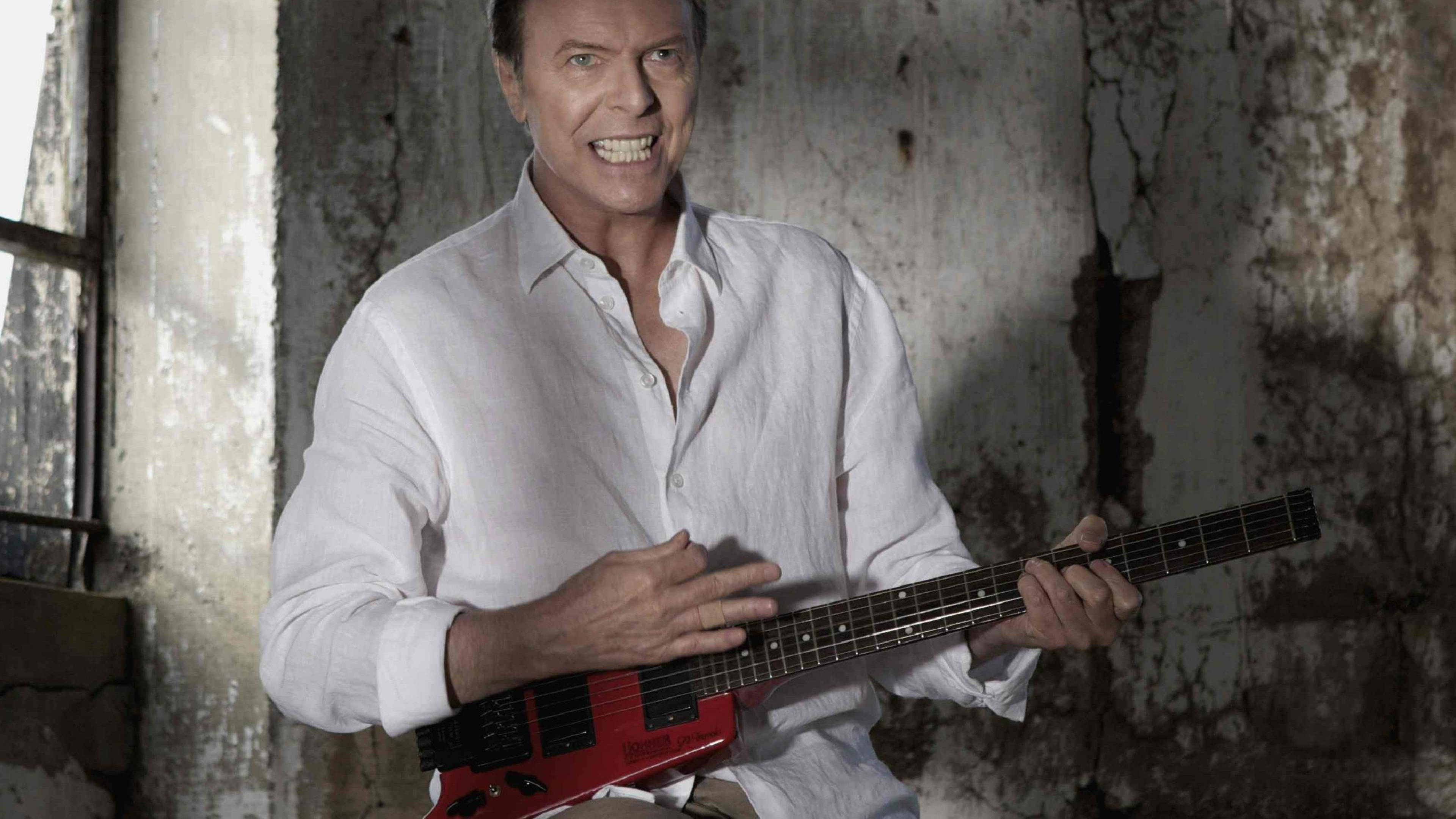 David Bowie 桌布 壁紙 — 大衛鮑伊咧嘴大笑，手持紅色電吉他，靠著風化的工業牆。