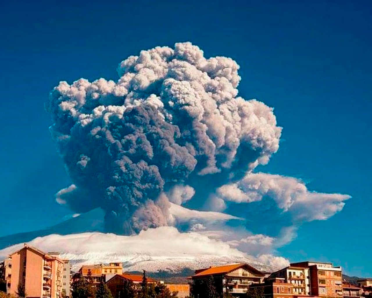Volcano 桌布 壁紙 — 大規模火山爆發將巨大的火山灰柱噴向蔚藍天空，下方是白雪覆蓋的山峰和附近的城鎮。