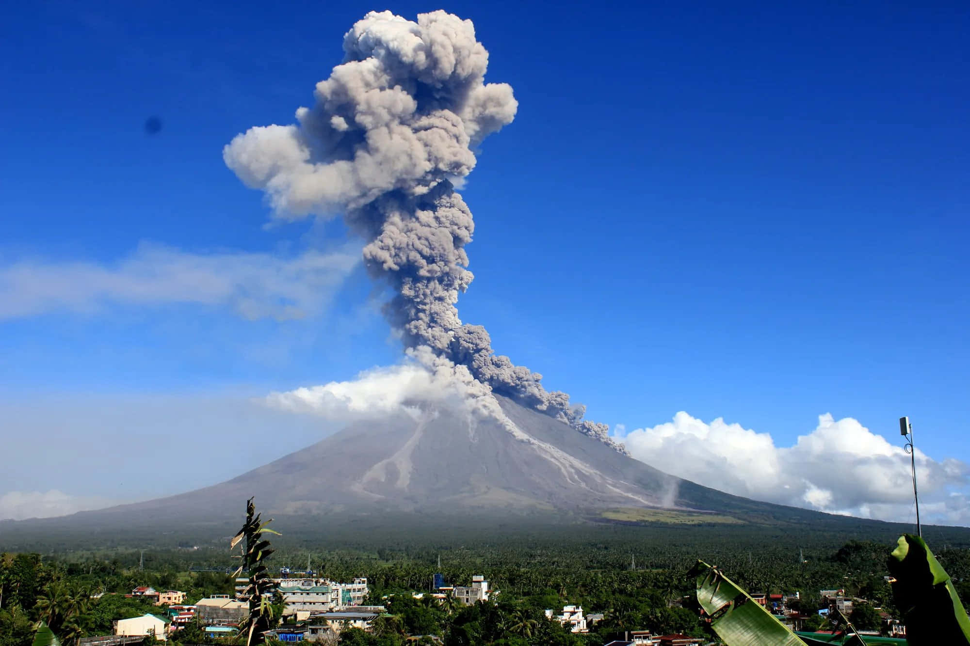 Natural Disaster 桌布 壁紙 — 大規模火山爆發將高聳的火山灰柱噴向蔚藍天空，下方是鬱鬱蔥蔥的綠色景觀和遠處的城鎮。