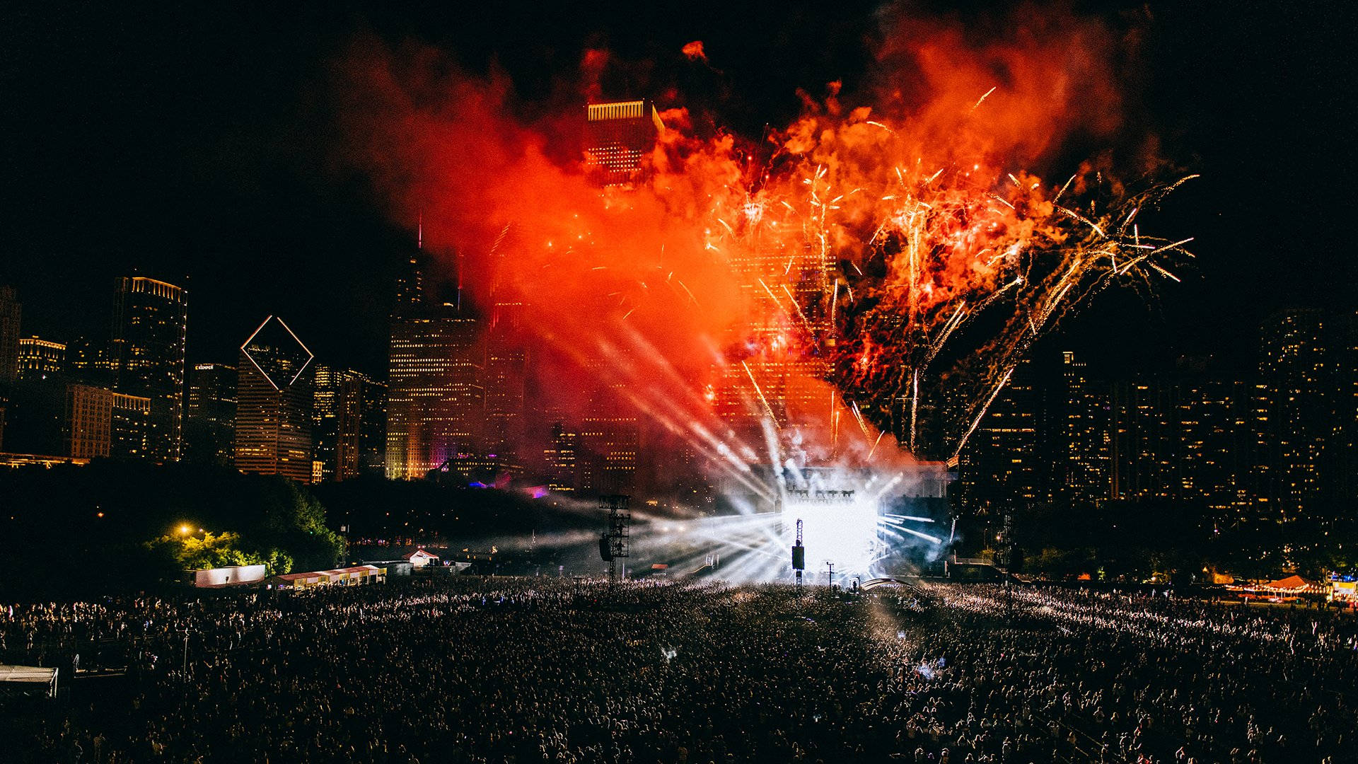 Lollapalooza 桌布 壁紙 — 大量人群在夜晚聚集參加音樂會，燦爛的紅色煙火在燈火通明的城市天際線上爆炸。