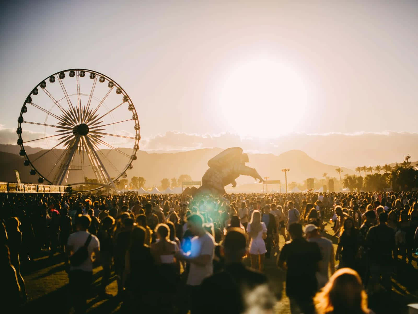Coachella 桌布 壁紙 — 大量人群聚集在節日活動中，太陽在巨大的摩天輪和山脈輪廓後落下。