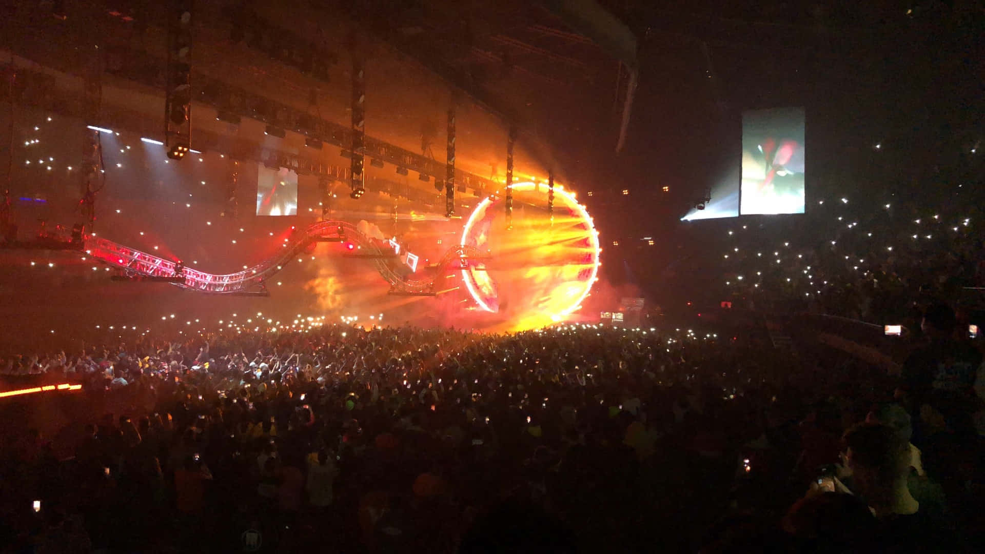 Travis Scott Concert 桌布 壁紙 — 大量演唱會觀眾在熾熱圓形舞台燈光和 towering 螢幕下舉起發光手機。