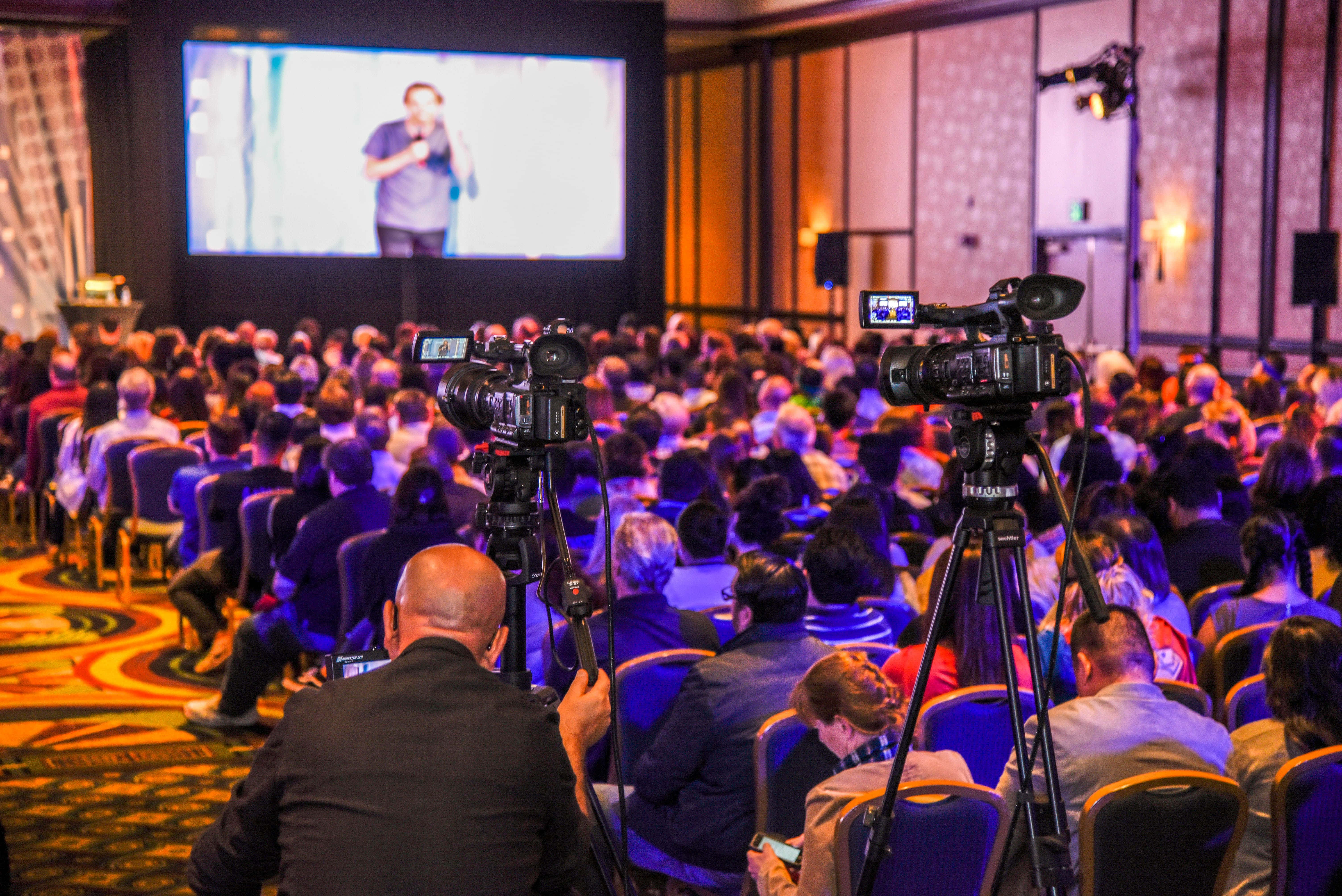 Corporate Event 桌布 壁紙 — 大量觀眾在溫暖的環境燈光下觀看台上講者，專業攝影機正在捕捉活動。