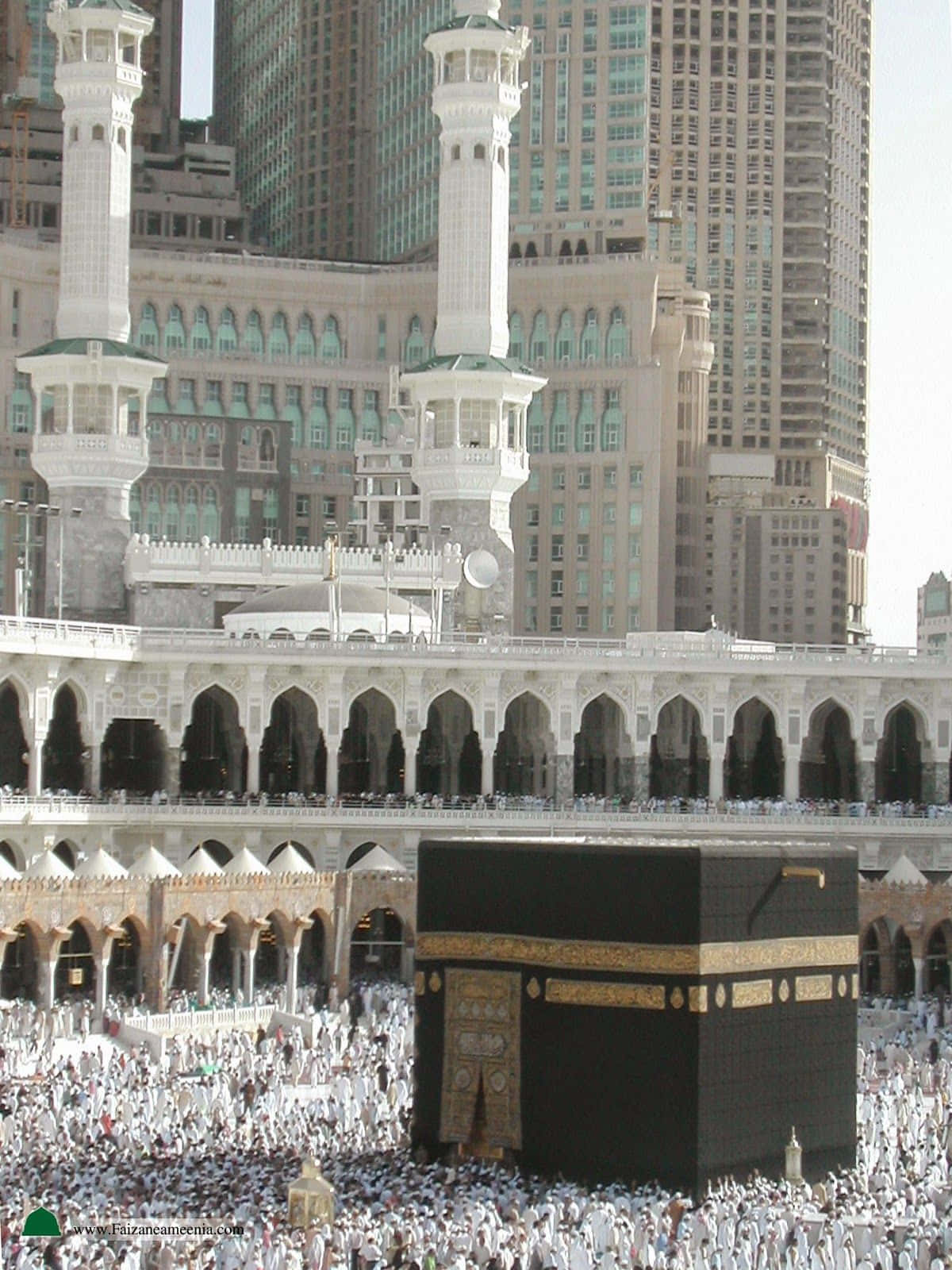 Makkah 桌布 壁紙 — 大量身穿白衣的人群在明亮的日光下圍繞著黑色天房，現代摩天大樓在裝飾華麗的白色宣禮塔後方高聳入雲。