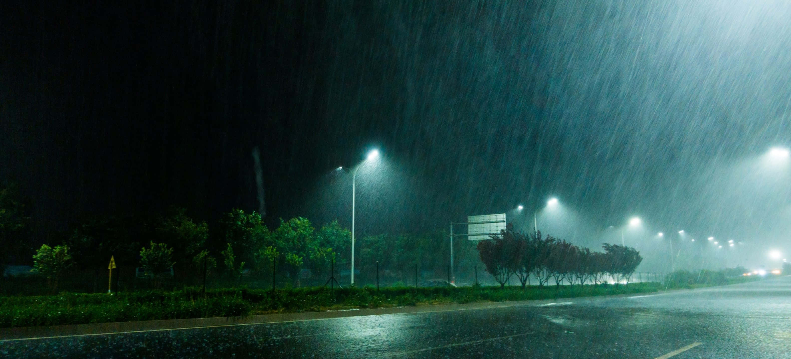 3168x1440 桌布 壁紙 — 大雨在夜晚沿著黑暗的城市街道傾瀉而下，被發光的街燈照亮，反射在潮濕的路面上。