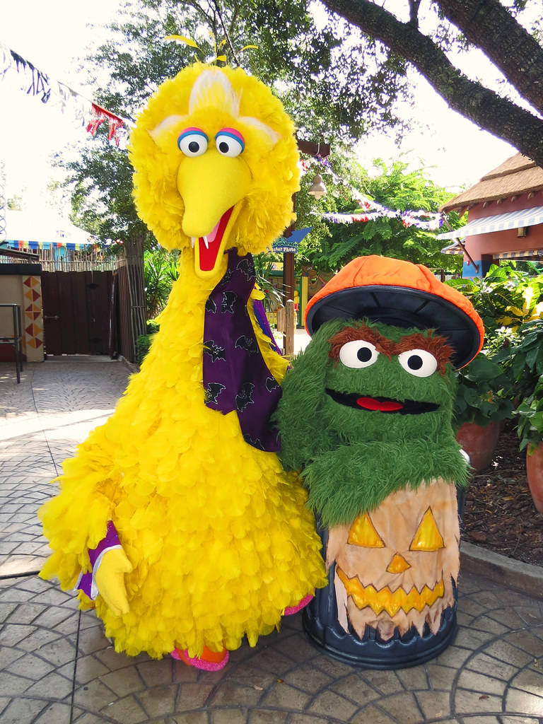 Big Bird 桌布 壁紙 — 大鳥和奧斯卡在萬聖節服裝中一起站在鋪砌的小路上，頭頂是枝葉茂密的樹木。