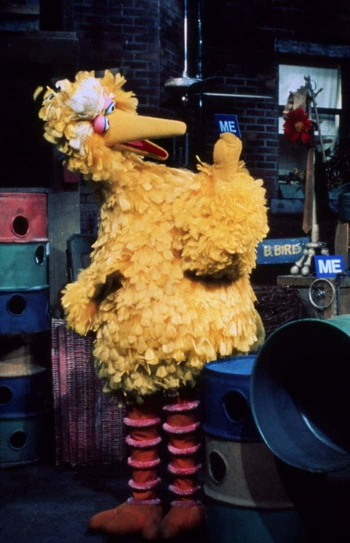 Big Bird 桌布 壁紙 — 大鳥在一個雜亂的工作坊中高高站立，身後有彩色桶和磚牆。