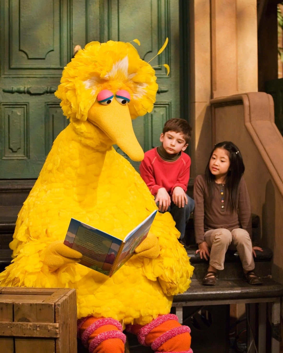 Big Bird 桌布 壁紙 — 大鳥讀一本書給兩個坐在舞台上的孩子，舞台上有綠色門和木箱。