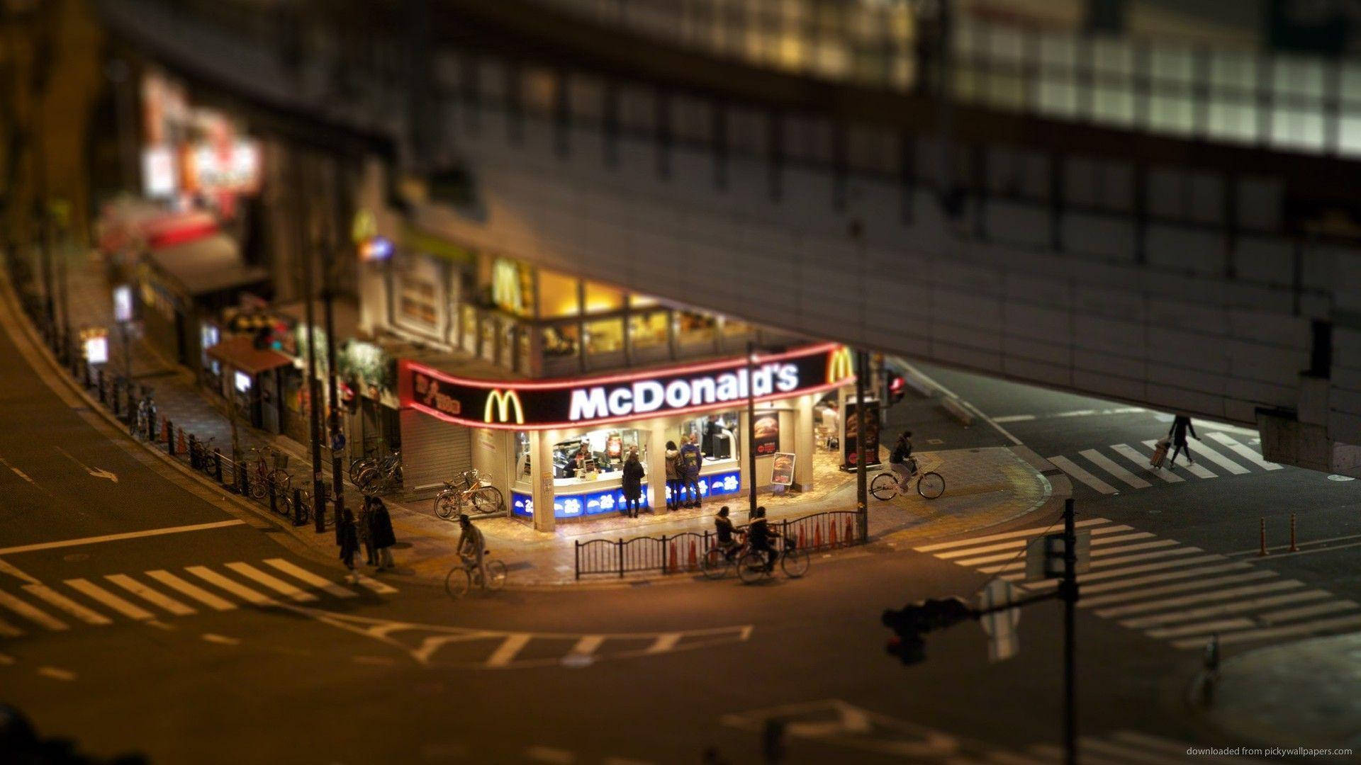 麥當勞 桌布 壁紙 — 天橋下亮燈的麥當勞，彎曲的城市街道上有行人和騎腳踏車人士的微型風格夜景。