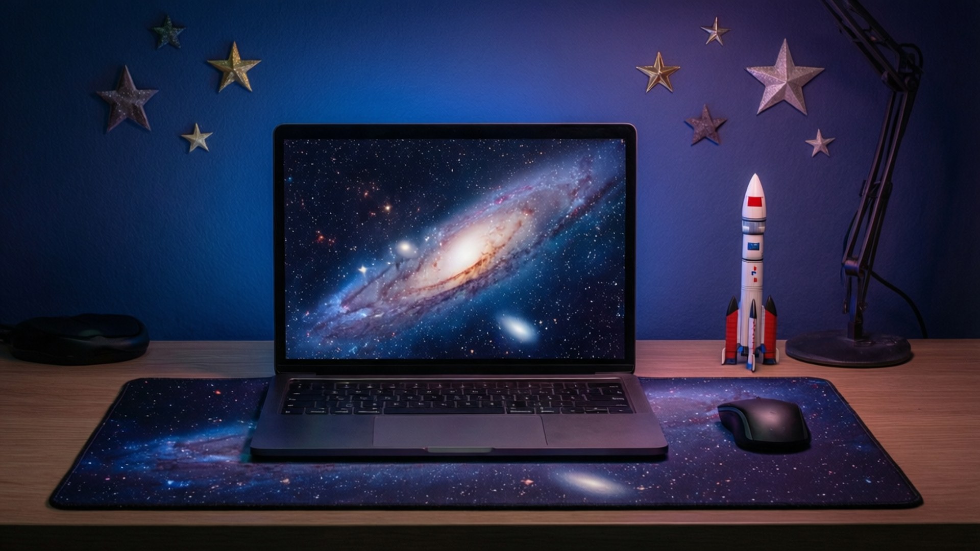 太空探險家桌面 壁紙 — 一台顯示著充滿活力的螺旋星系的筆記型電腦，放在一個星形滑鼠墊上，旁邊有一個玩具火箭和檯燈，背景是藍色牆壁，上面裝飾著金屬星星。