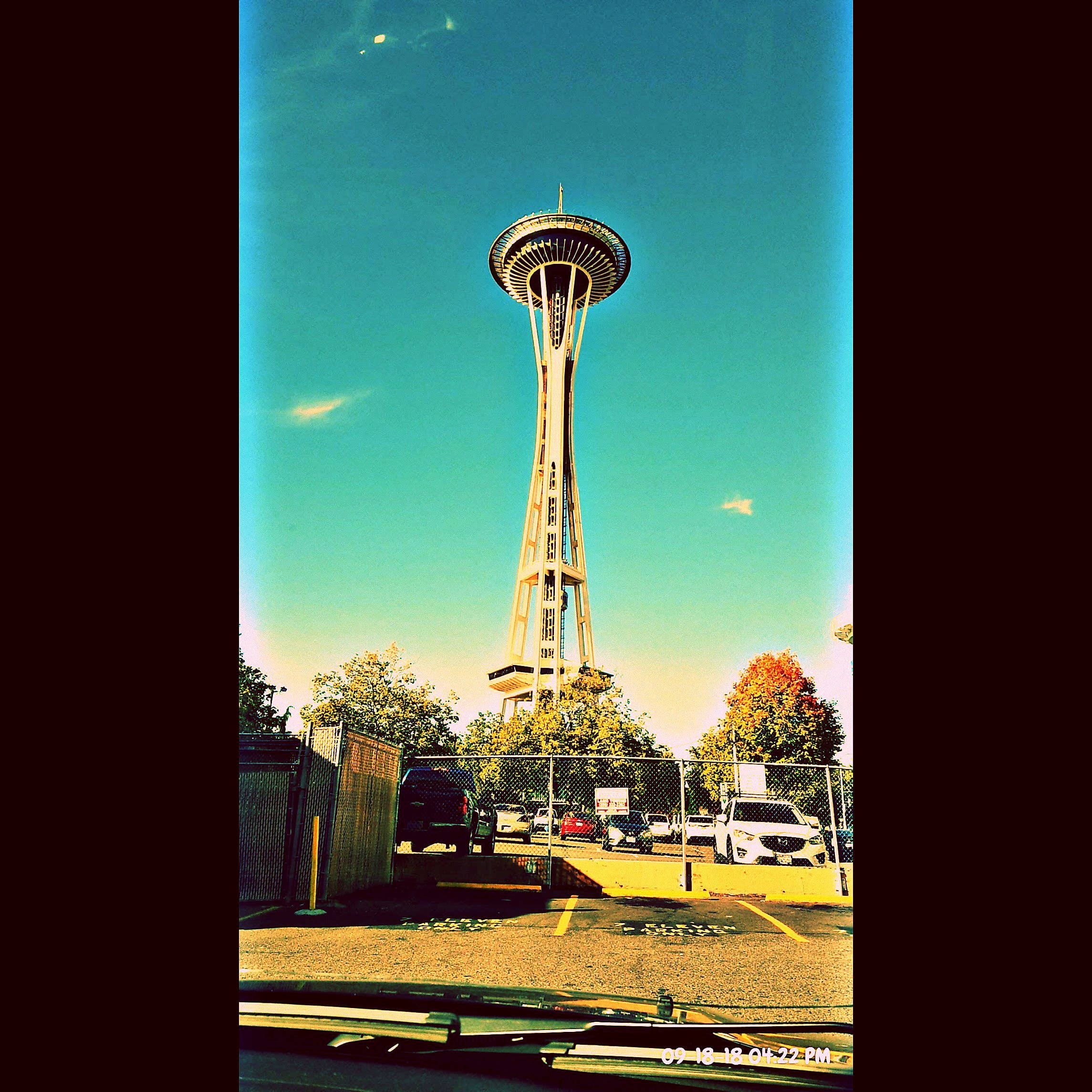 Space Needle 桌布 壁紙 — 太空針塔在充滿活力的藍綠色天空下，聳立於一個停車場之上，下方有秋天的樹木和汽車。