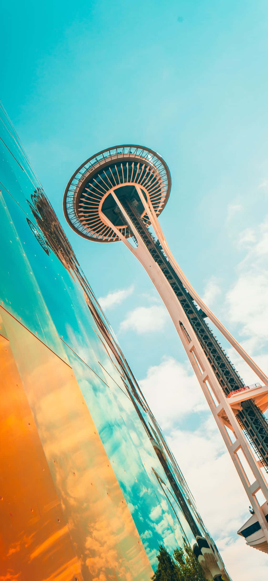 iPhone X Seattle 桌布 壁紙 — 太空針塔在明亮的藍天下高聳，其倒影在色彩繽紛的玻璃建築外牆上閃爍。