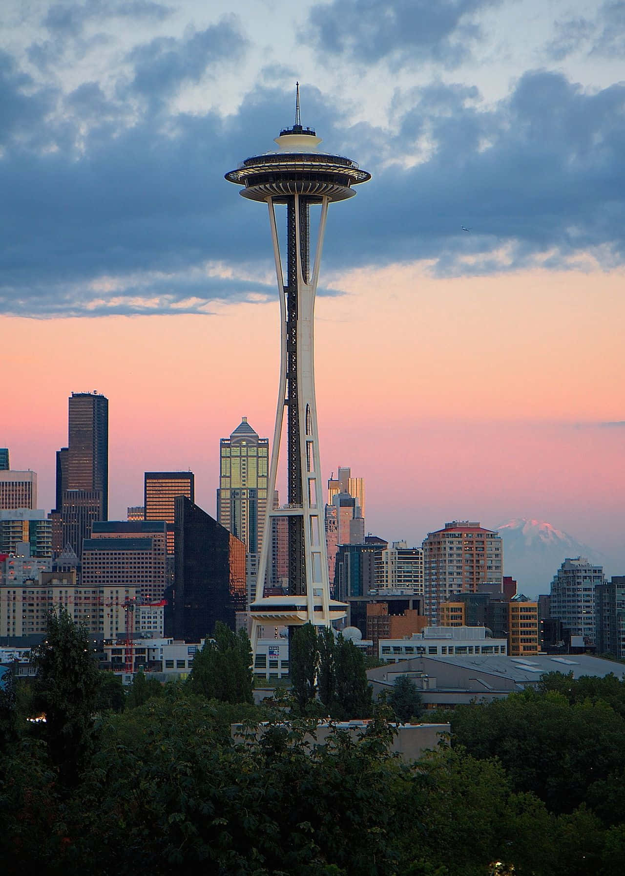 Best Seattle 桌布 壁紙 — 太空針塔在黃昏時分聳立於西雅圖天際線之上，上方有粉紅色和藍色的雲朵，前景有綠色樹木。