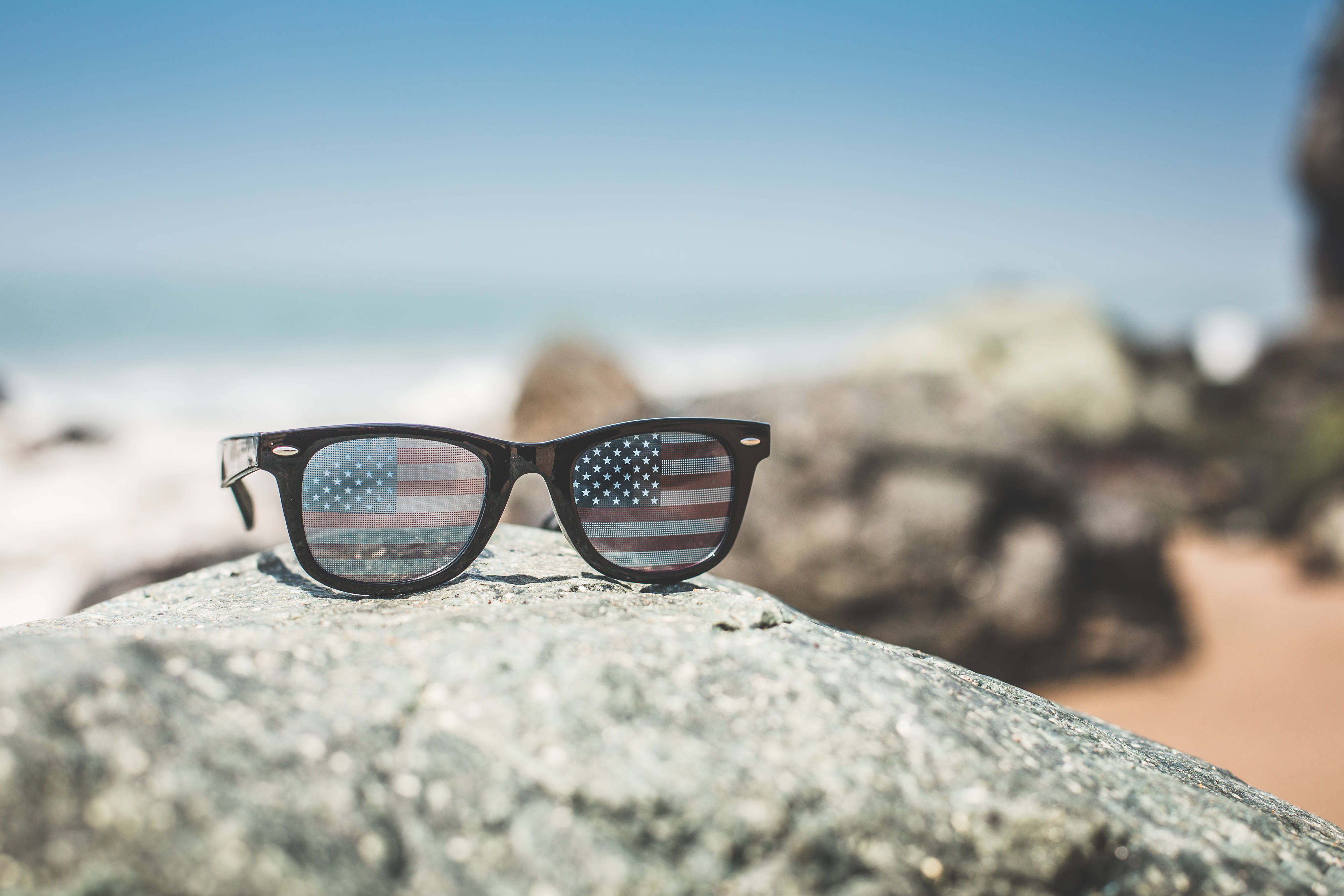 USA flag iPhone 桌布 壁紙 — 太陽眼鏡上反射出美國國旗，擱在陽光明媚的海岸沙灘岩石上，背景是清澈的藍天。