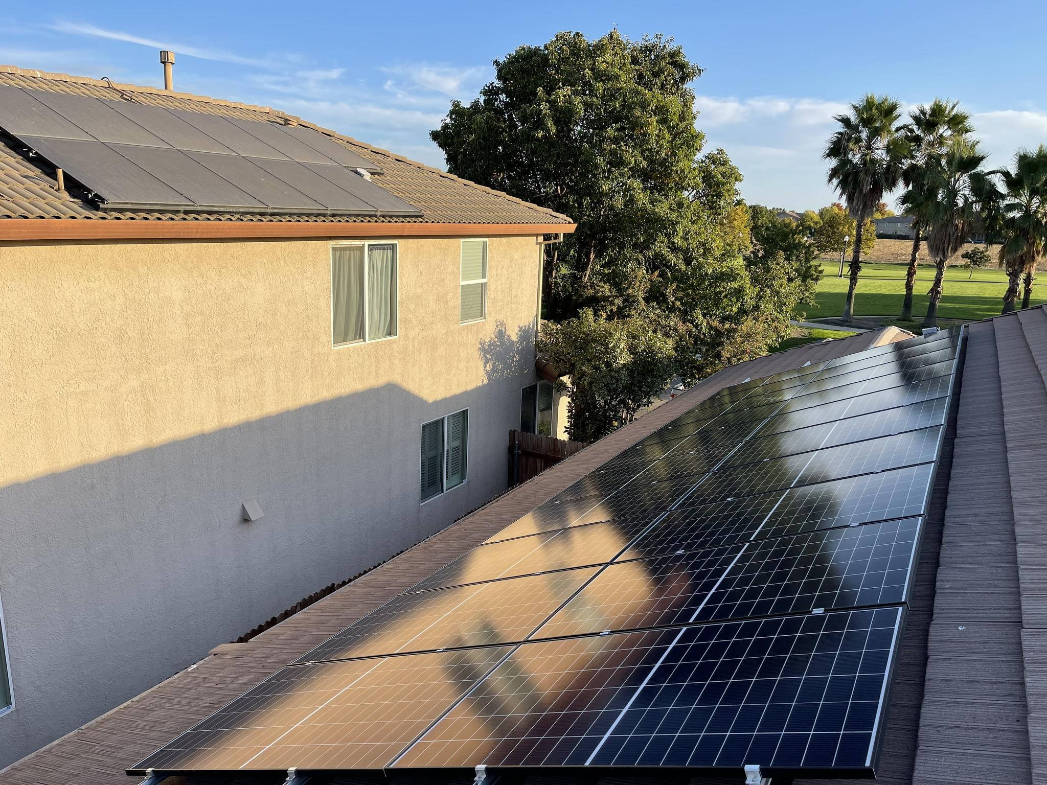 Solar Panel 桌布 壁紙 — 太陽能板在晴朗藍天下，以棕櫚樹和綠色草坪為背景，在住宅屋頂上閃閃發光。