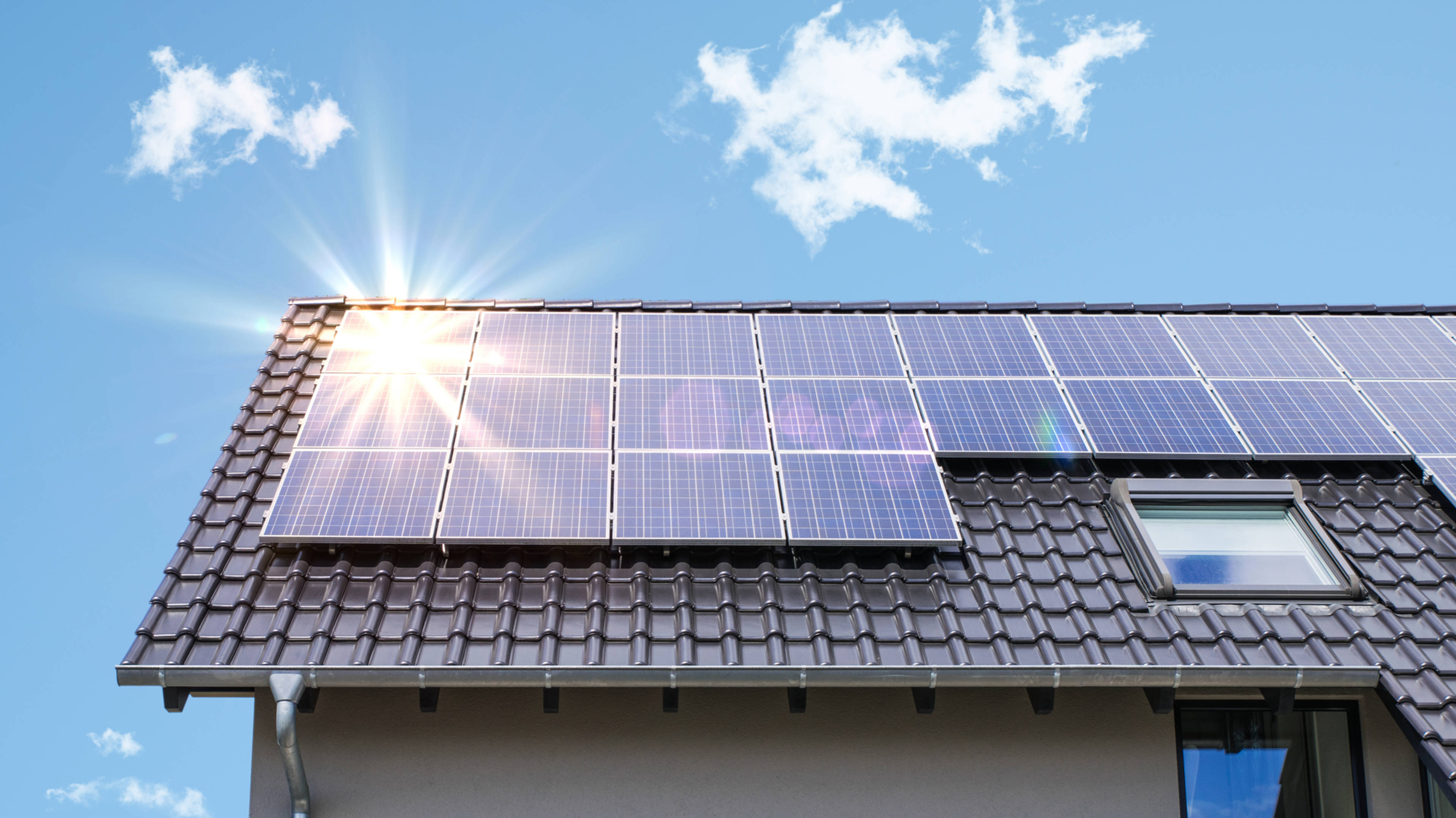 Solar Panel 桌布 壁紙 — 太陽能電池板在明亮的陽光下在瓦片屋頂上閃閃發光，背景是清澈的藍天和縷縷白雲。