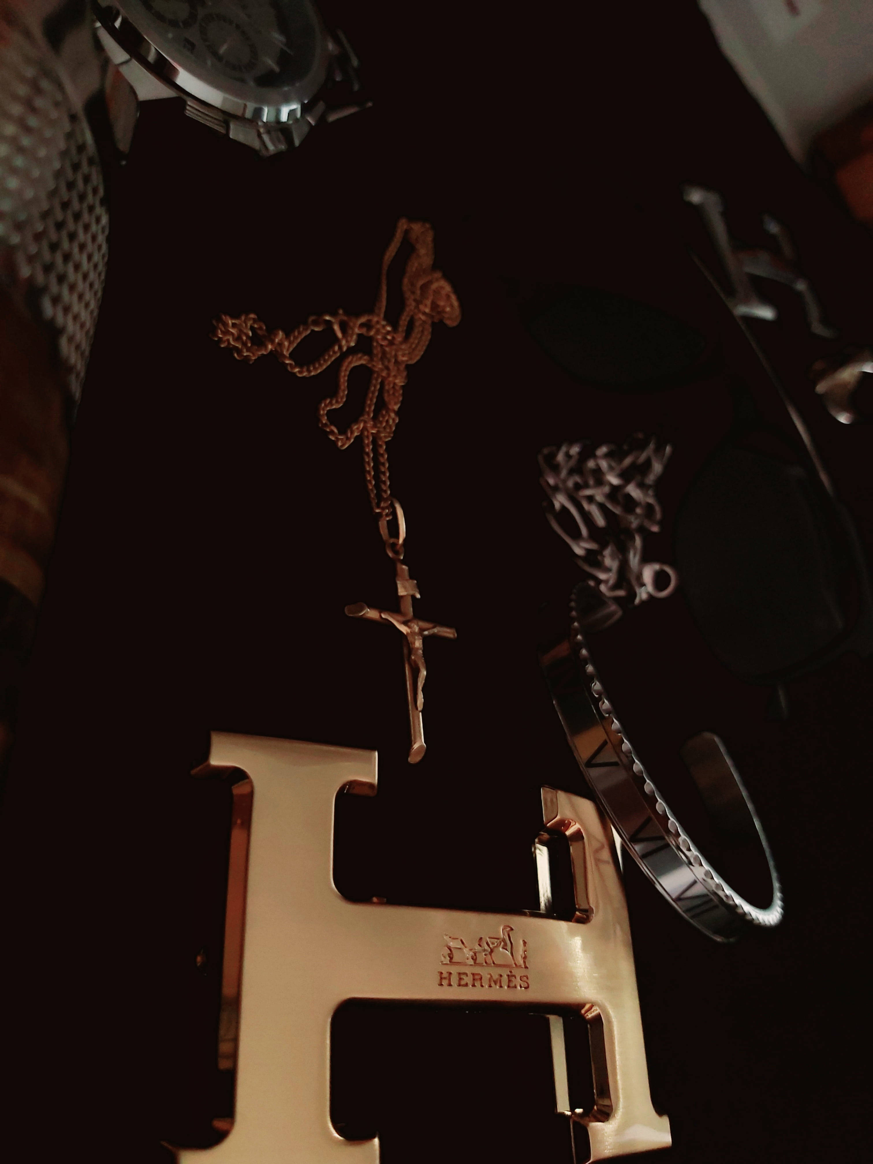 Hermes 桌布 壁紙 — 奢華配件，包括金色十字項鍊、Hermès皮帶扣和銀色手錶，排列在深色表面上。
