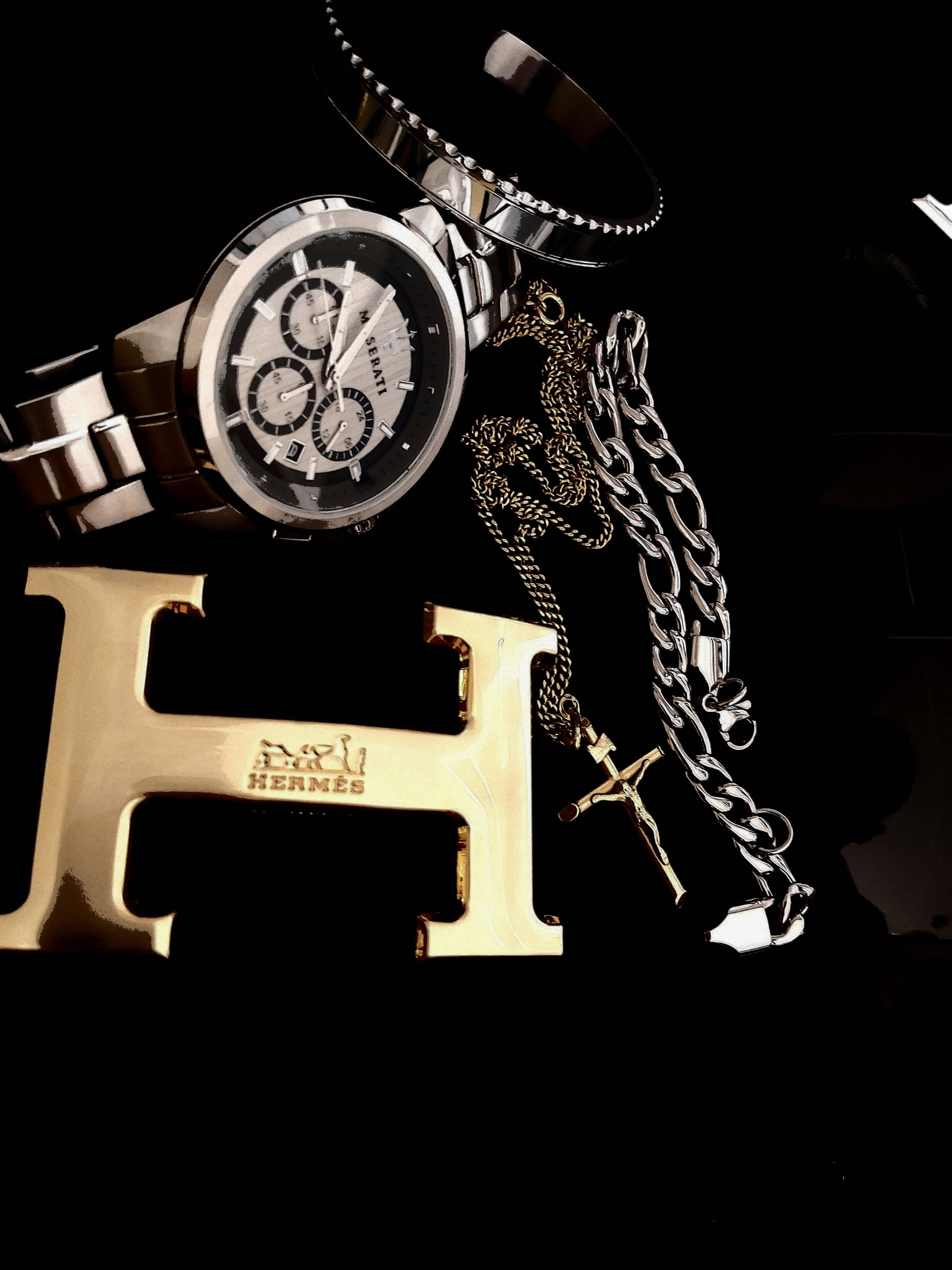 Hermes 桌布 壁紙 — 奢華配件，包括銀色計時碼錶、金色愛馬仕皮帶扣和粗鏈條，在純黑背景下閃耀。