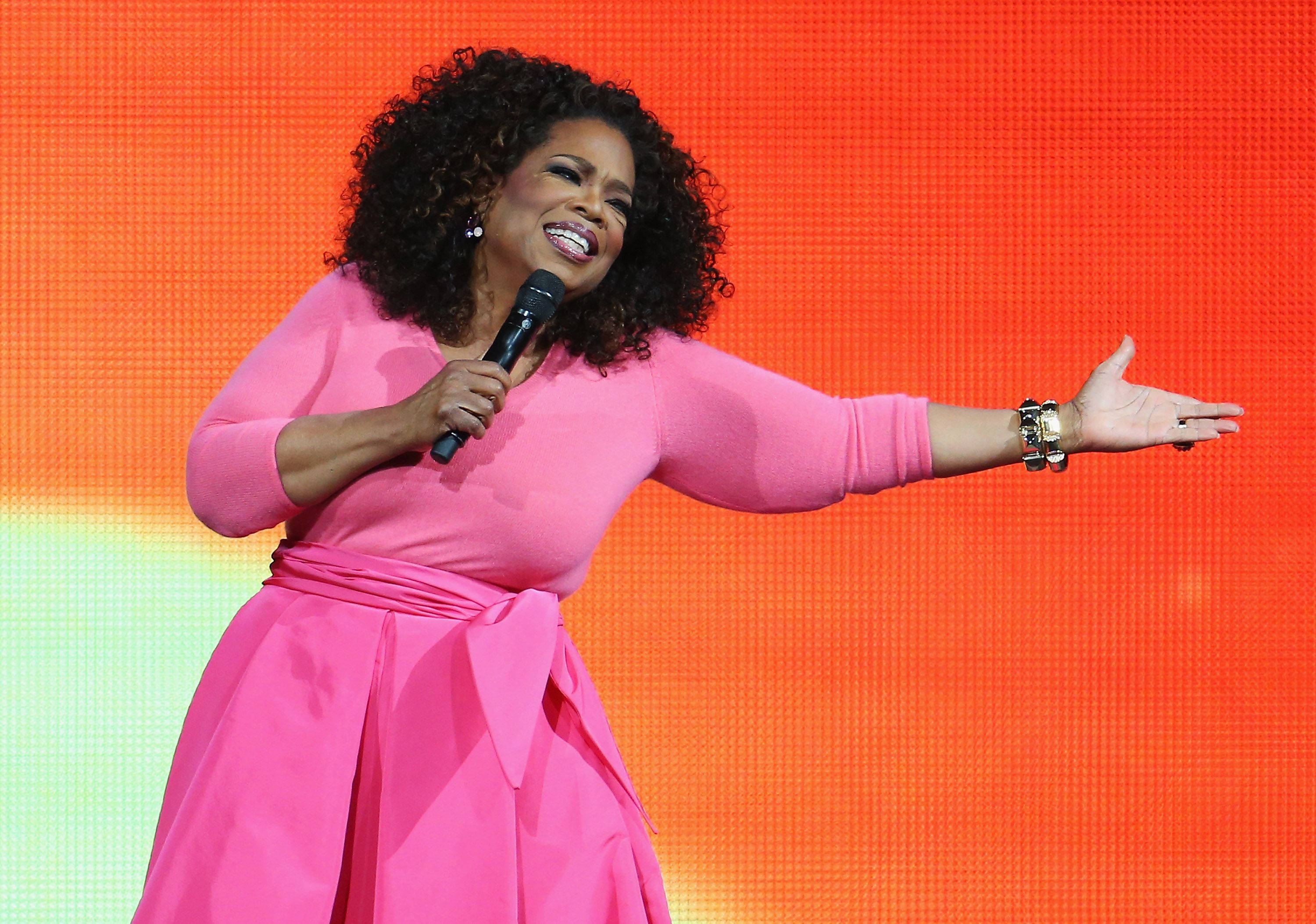 Oprah 桌布 壁紙 — 奧普拉·溫芙瑞在充滿活力的橙綠色漸變舞台上對著麥克風溫暖地微笑說話。