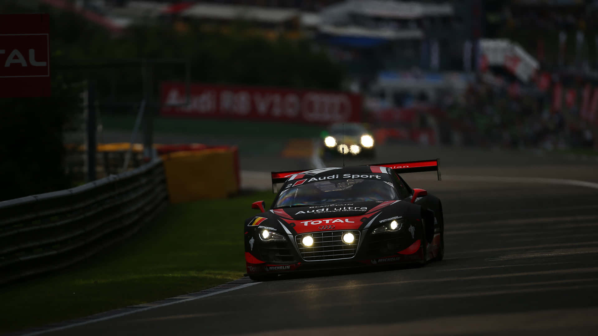 Racing 桌布 壁紙 — 奧迪賽車閃爍車頭燈，在昏暗的夜間賽道上疾馳，背景有模糊的觀眾和贊助商橫幅。