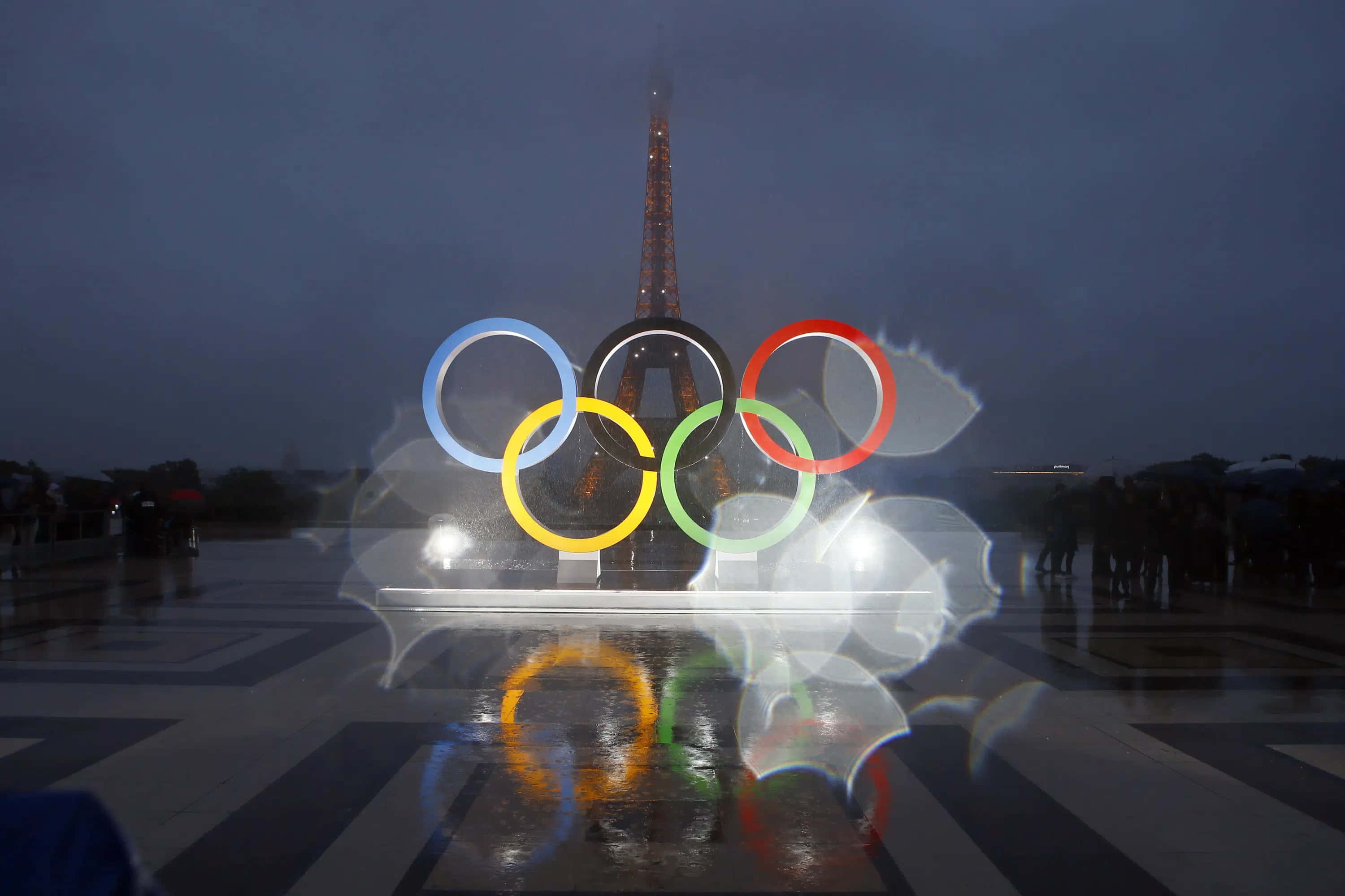 Olympics 桌布 壁紙 — 奧運五環在雨夜天空中閃耀，背後艾菲爾鐵塔高聳矗立。