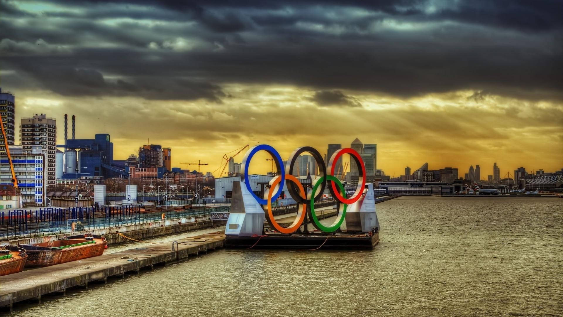 Olympics 桌布 壁紙 — 奧運五環漂浮在水面上，背景是戲劇性的金色日落雲彩和城市天際線。