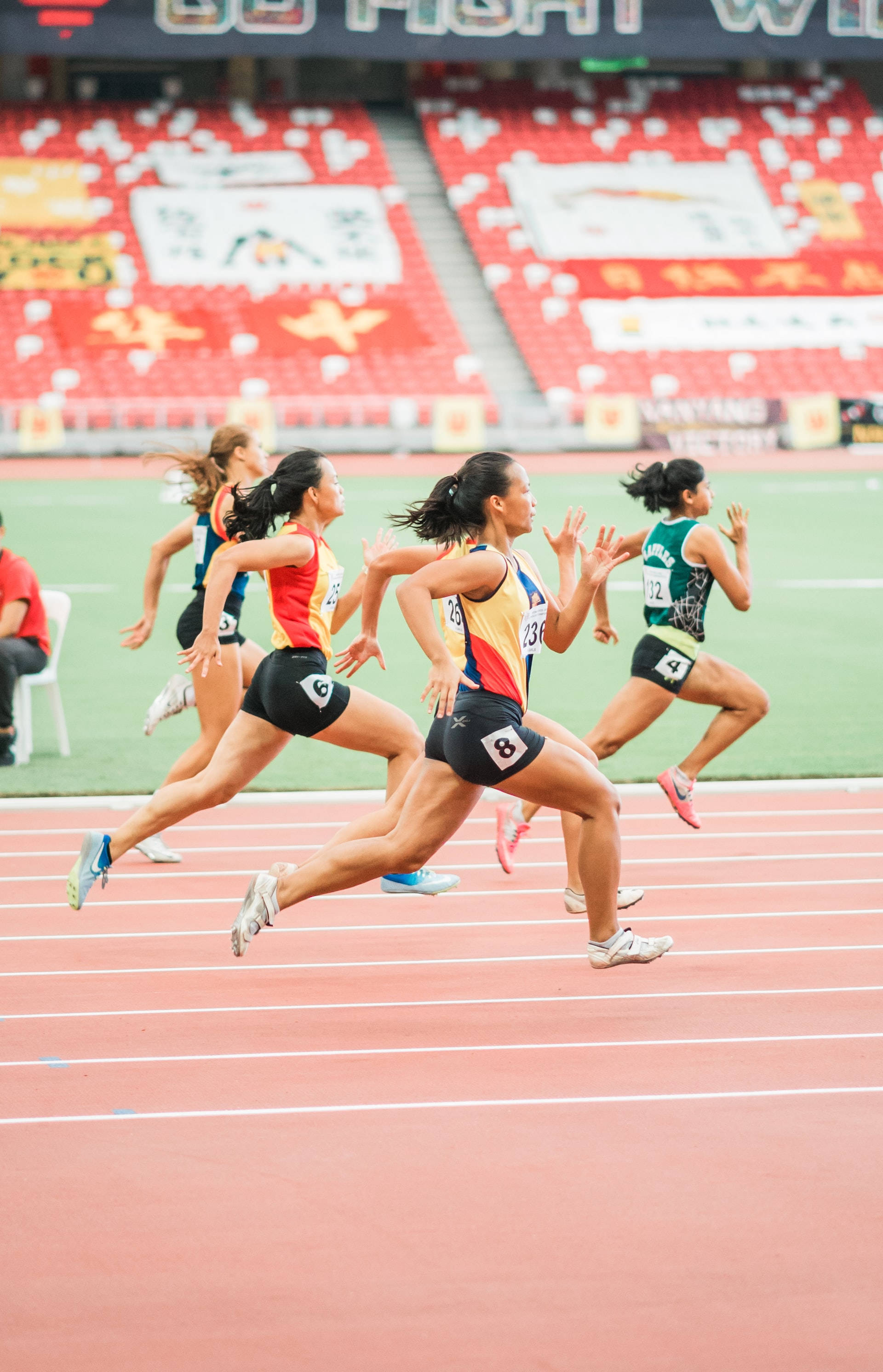 Athletic 桌布 壁紙 — 女性短跑選手在紅色跑道上激烈比賽，背景中的體育場座位模糊不清。