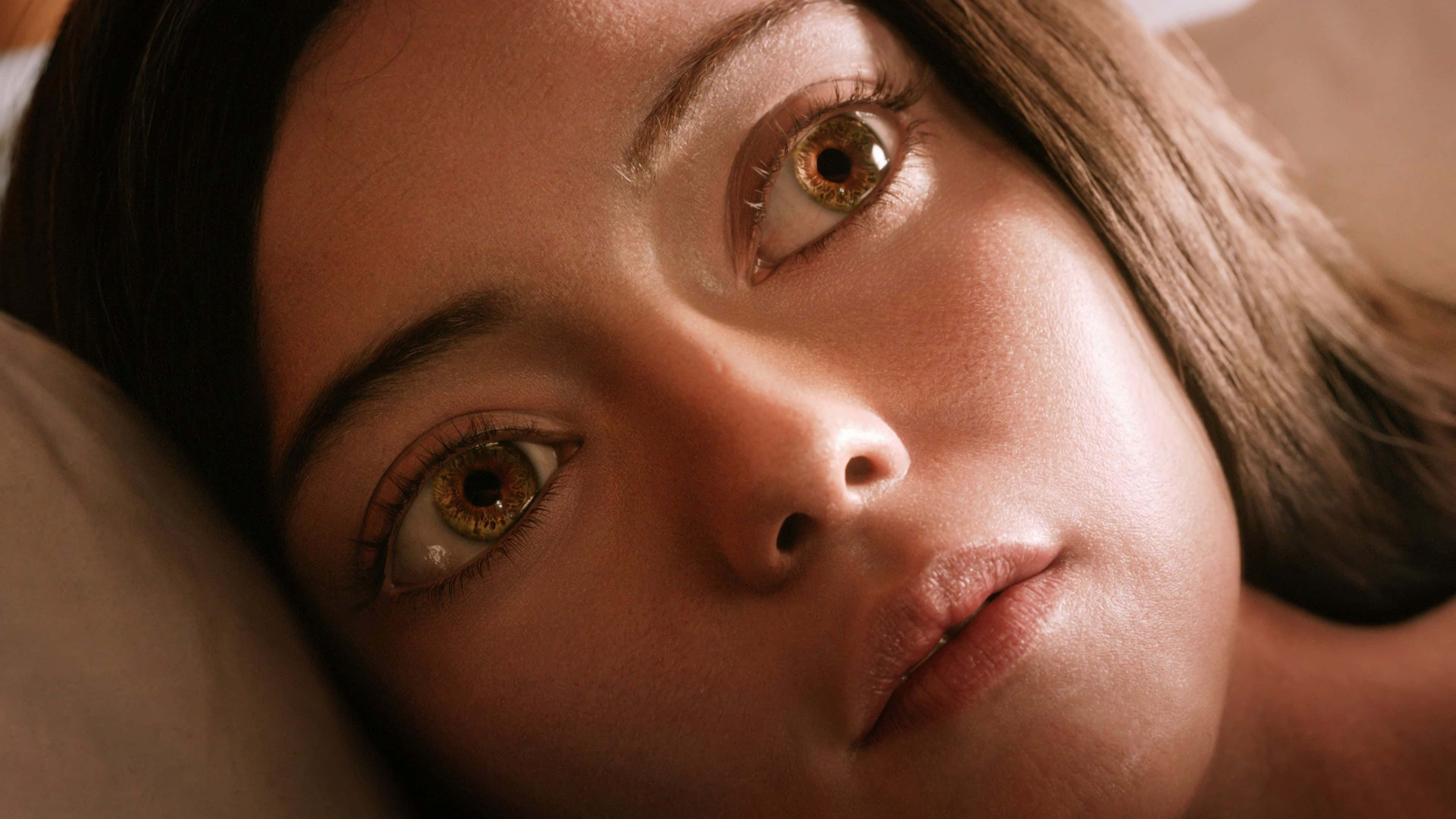 Alita Battle Angel 桌布 壁紙 — 女性臉部特寫，靠在枕頭上，溫暖的燈光突顯她細緻的棕色眼睛和柔軟的皮膚質感。