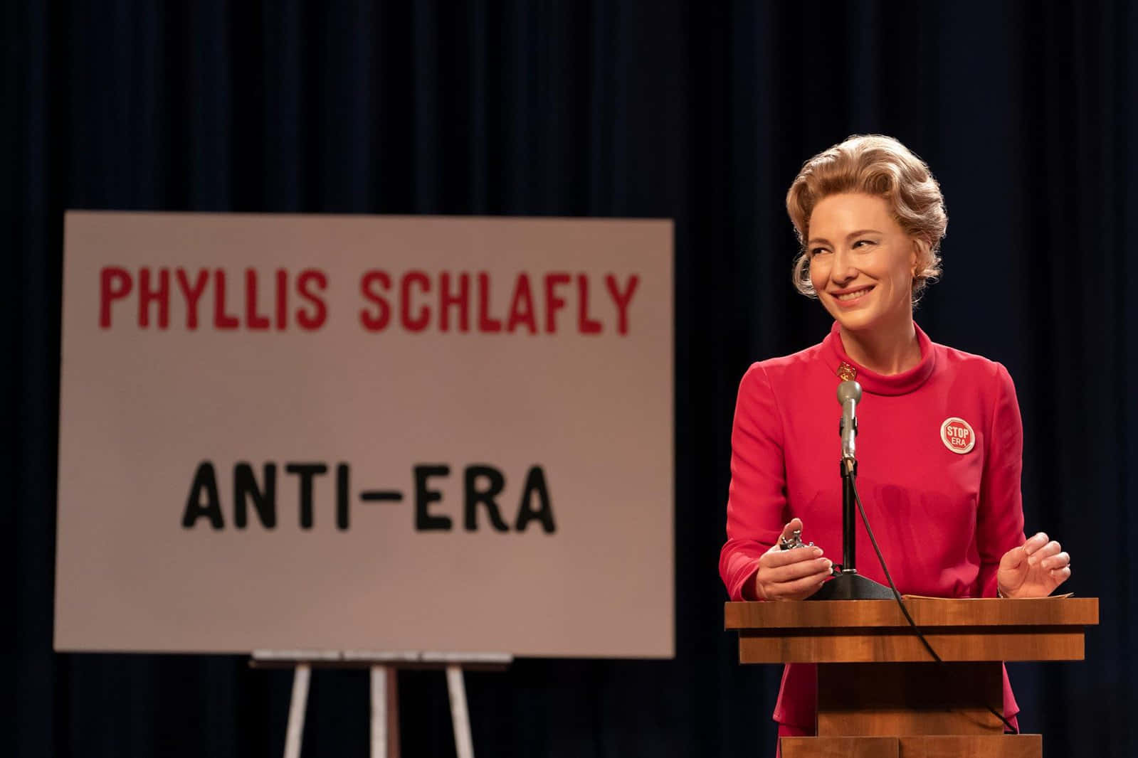 Cate Blanchett 桌布 壁紙 — 女演員凱特布蘭琪飾演菲莉絲施拉夫利，身穿紅色西裝，站在木製講台前，身後有標語牌寫著「PHYLIS SCHLAFLY ANTI-ERA」。