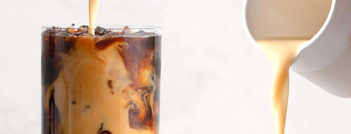 Milk And Coffee 桌布 壁紙 — 奶油在柔和白色背景上戲劇性地旋轉，倒入一杯加冰的冰咖啡中。