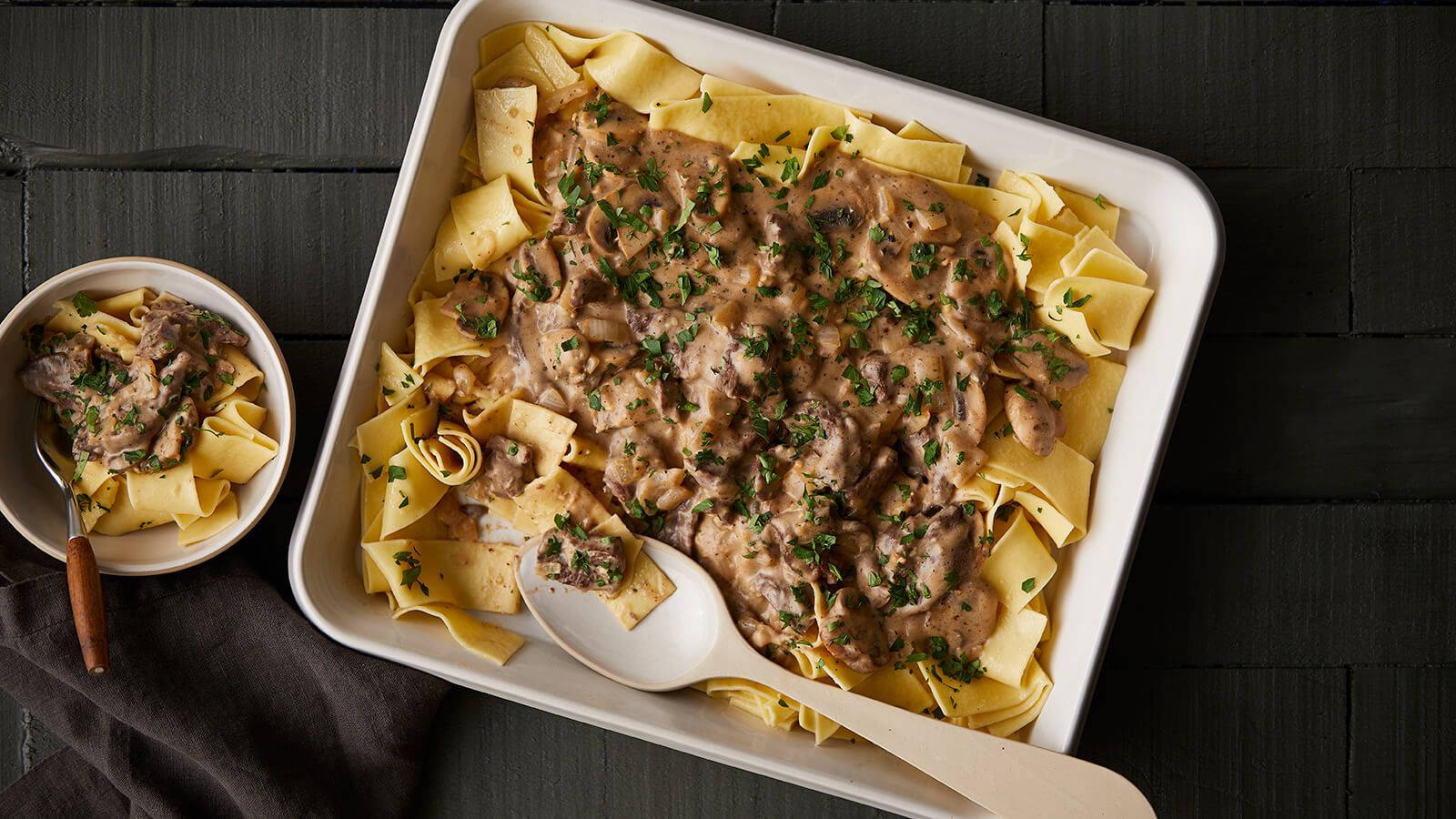 Pasta 桌布 壁紙 — 奶油牛肉斯特羅加諾夫搭配寬麵條，以新鮮歐芹點綴，盛放在白色烤盤中，置於深色木質表面上。