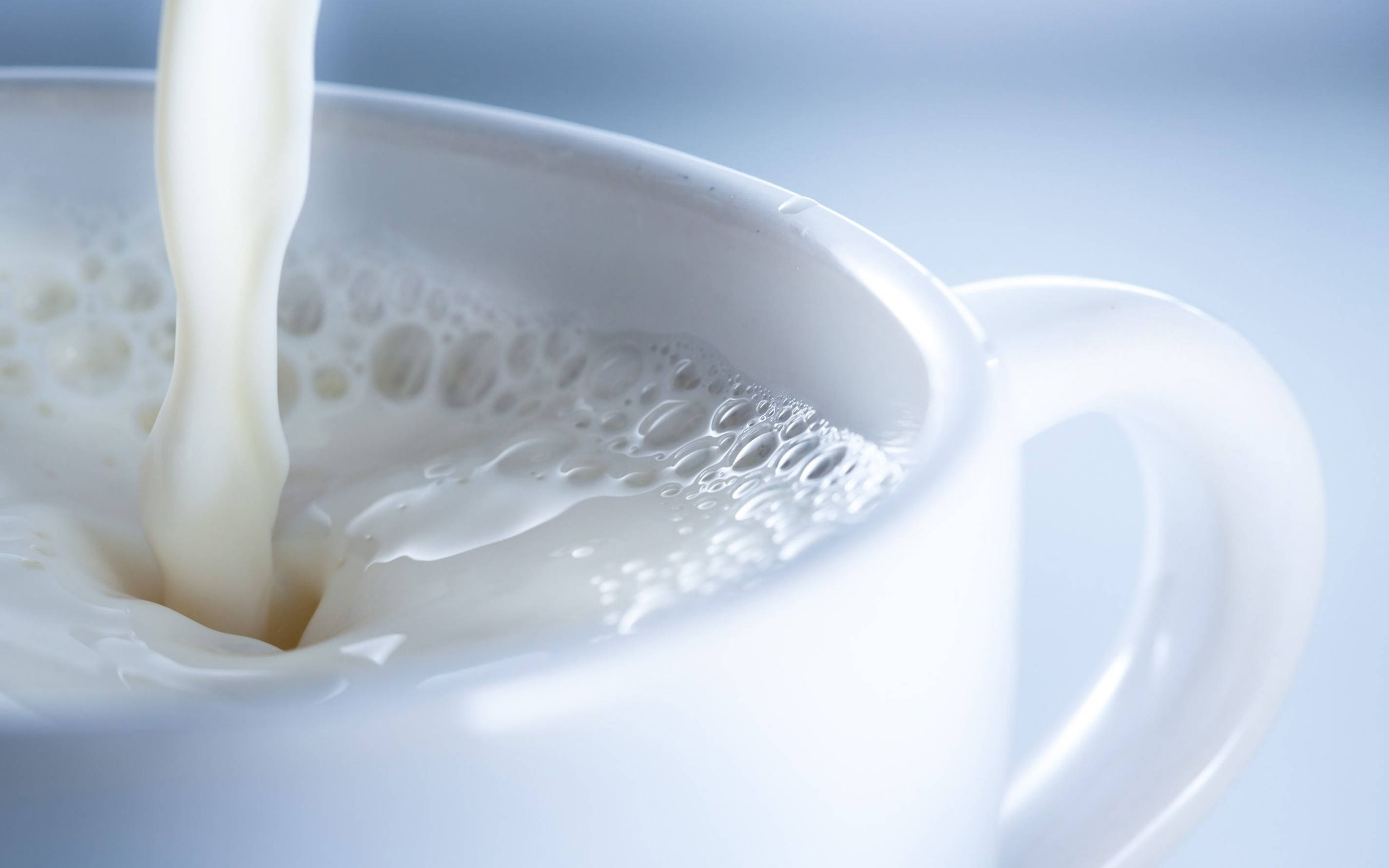 Milk 桌布 壁紙 — 奶油狀牛奶倒入白色陶瓷杯中，在柔和的藍色背景上形成旋轉的氣泡。