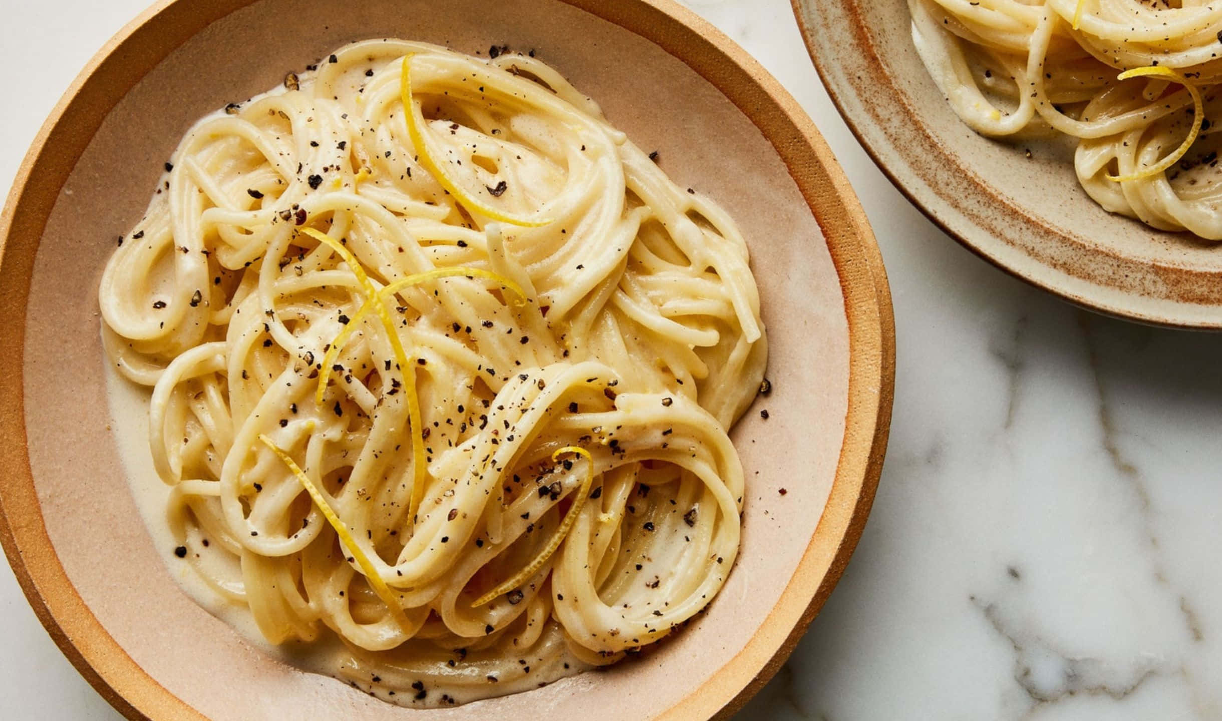 2440x1440 Pasta 桌布 壁紙 — 奶油義大利麵搭配檸檬皮和黑胡椒，盛裝在鄉村風木碗中，置於大理石檯面上。