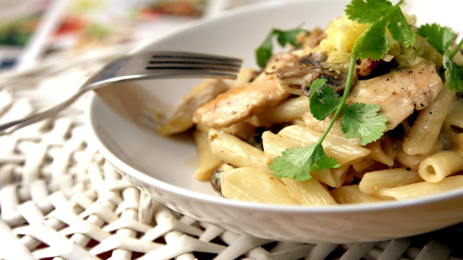 1920x1080 Pasta 桌布 壁紙 — 奶油雞肉筆管麵特寫，以新鮮香菜點綴，放在白色碗中，置於編織餐墊上。