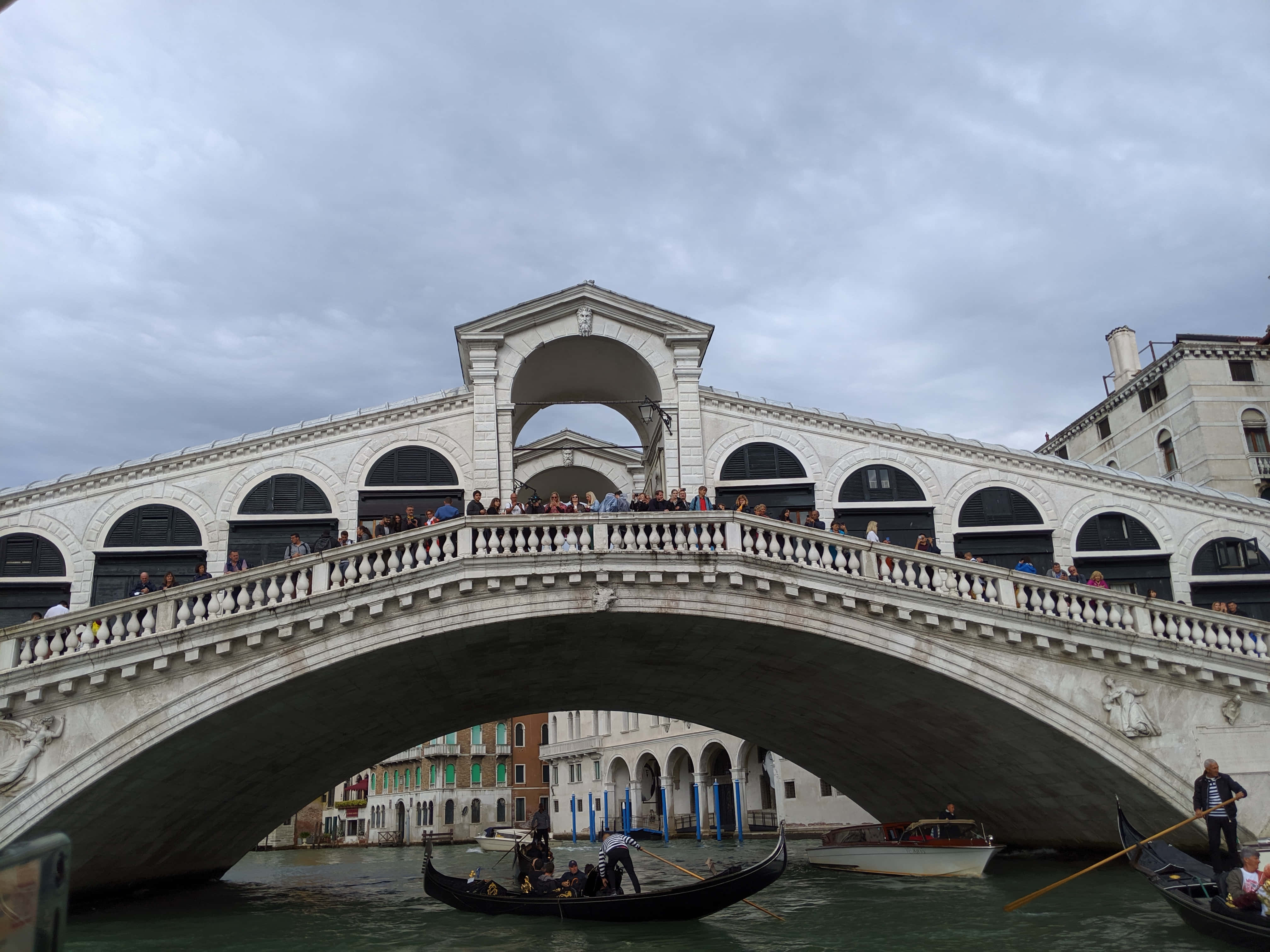 venice 桌布 壁紙 — 威尼斯標誌性的石造嘆息橋拱跨運河，下方有貢多拉滑行，遊客擠滿步道，天空多雲。