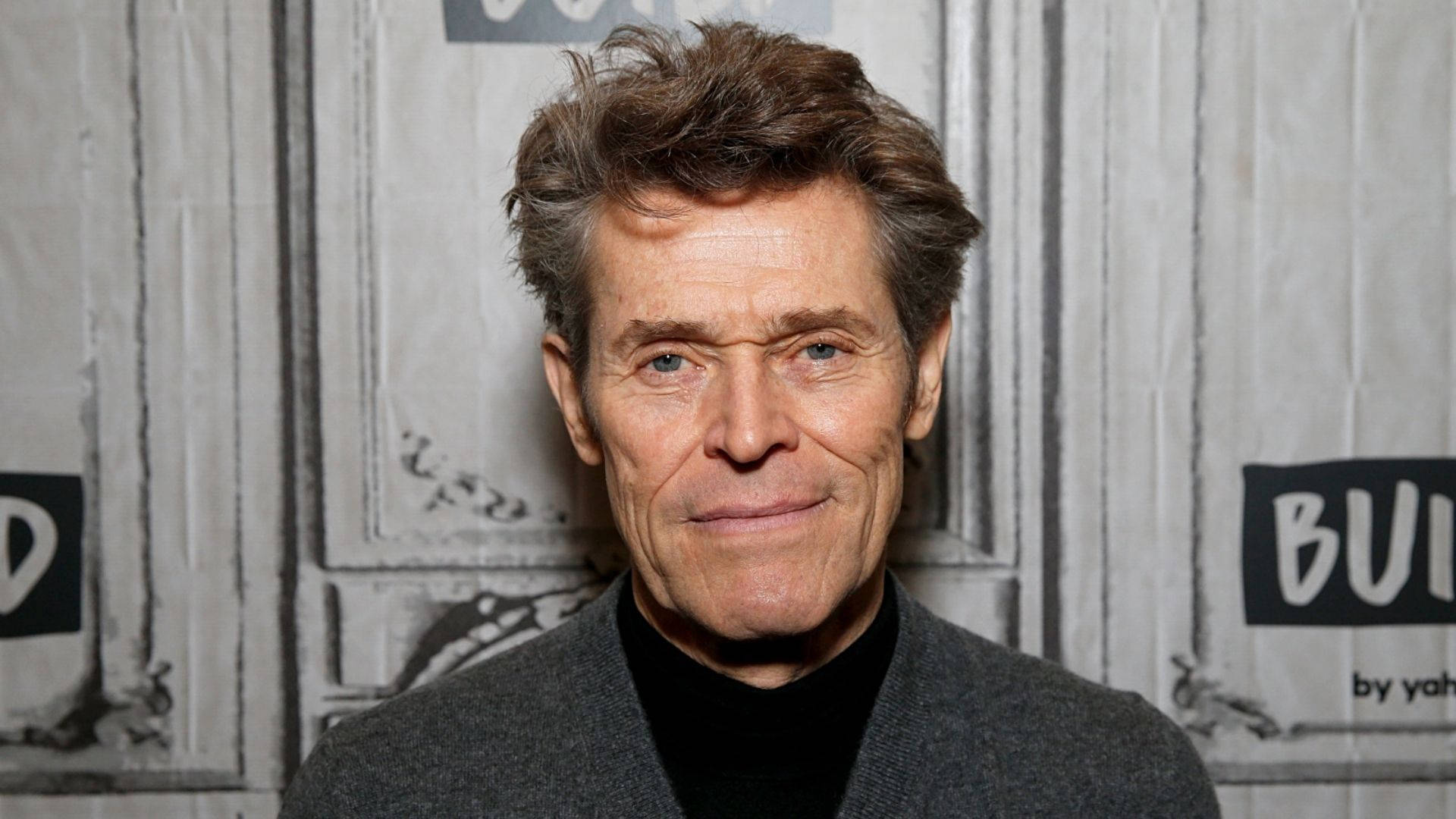 Willem Dafoe 桌布 壁紙 — 威廉·H·梅西的頭像特寫，一頭凌亂的棕色頭髮，臉上掛著含蓄的微笑，背景是帶有紋理的灰色背景，並有部分文字。