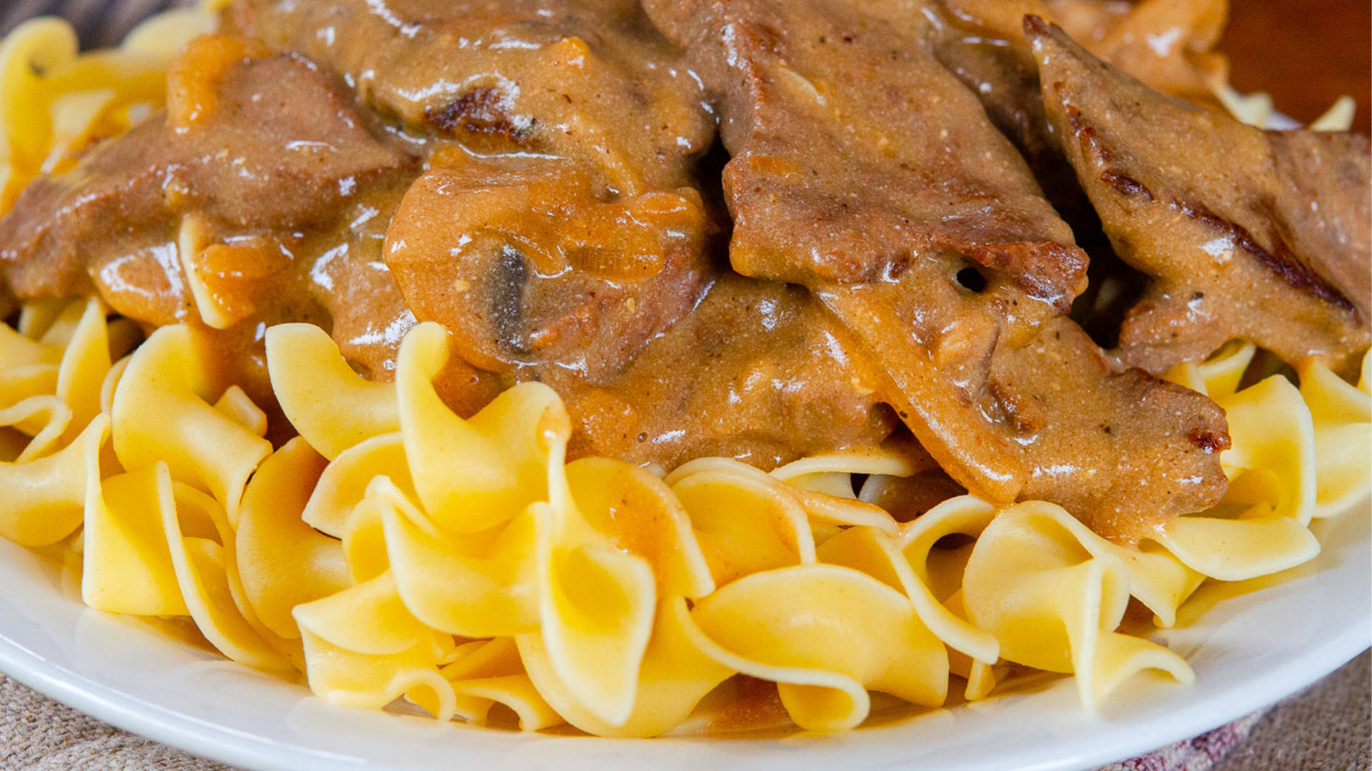 Pasta 桌布 壁紙 — 嫩滑牛肉燉菜搭配蘑菇和洋蔥，放在金色雞蛋麵上，盛於白色盤中的特寫。