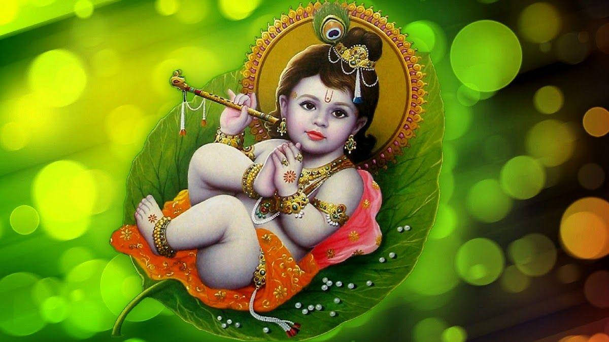 Little Krishna 桌布 壁紙 — 嬰兒克里希納盤腿坐在一片大綠葉上，吹奏著飾有珠子和寶石的長笛，背景是充滿活力的綠色散景。