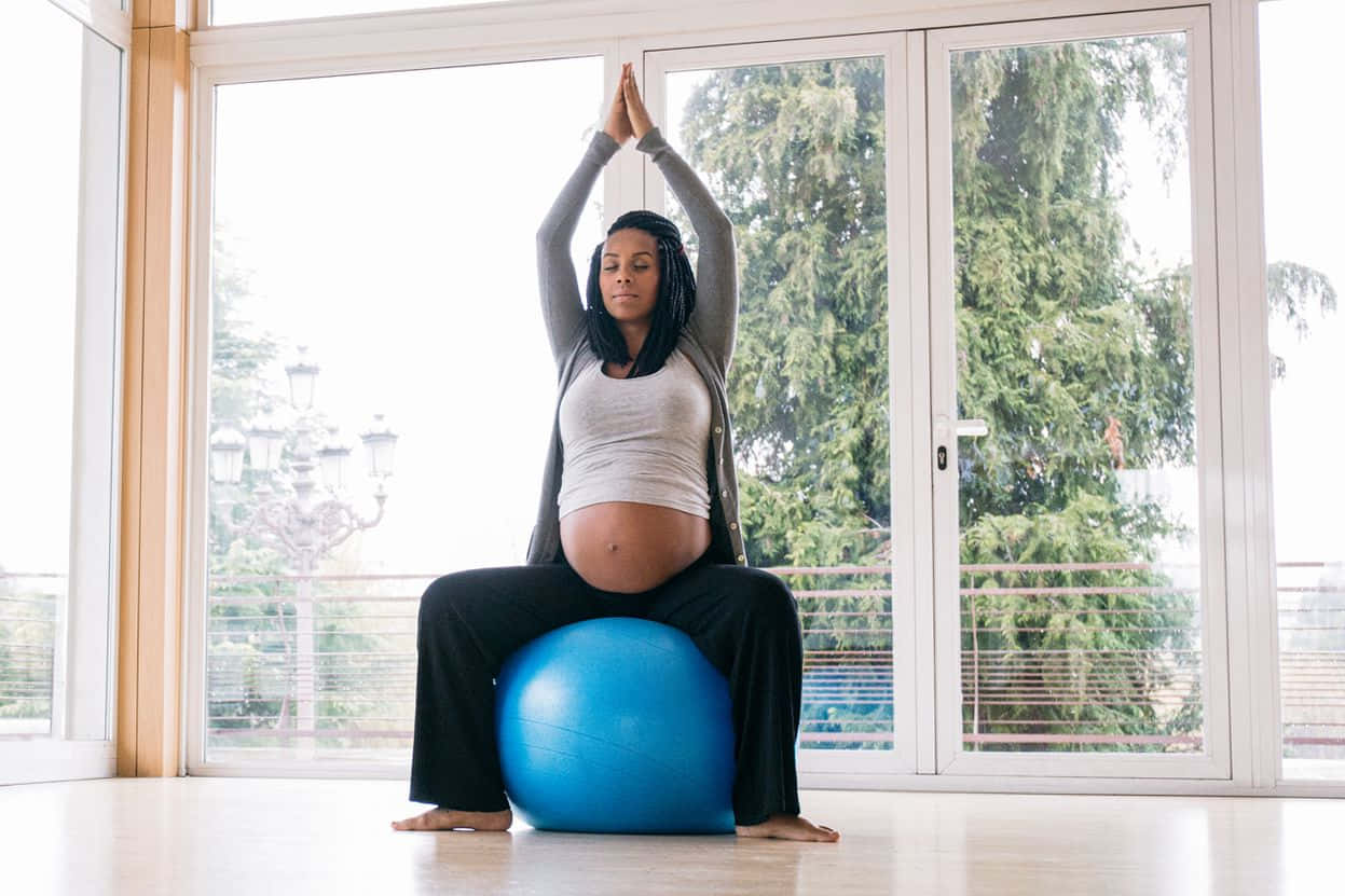 Exercise 桌布 壁紙 — 孕婦在室內藍色健身球上做出寧靜的瑜伽姿勢，透過大窗戶可以看到茂密的綠色樹木。