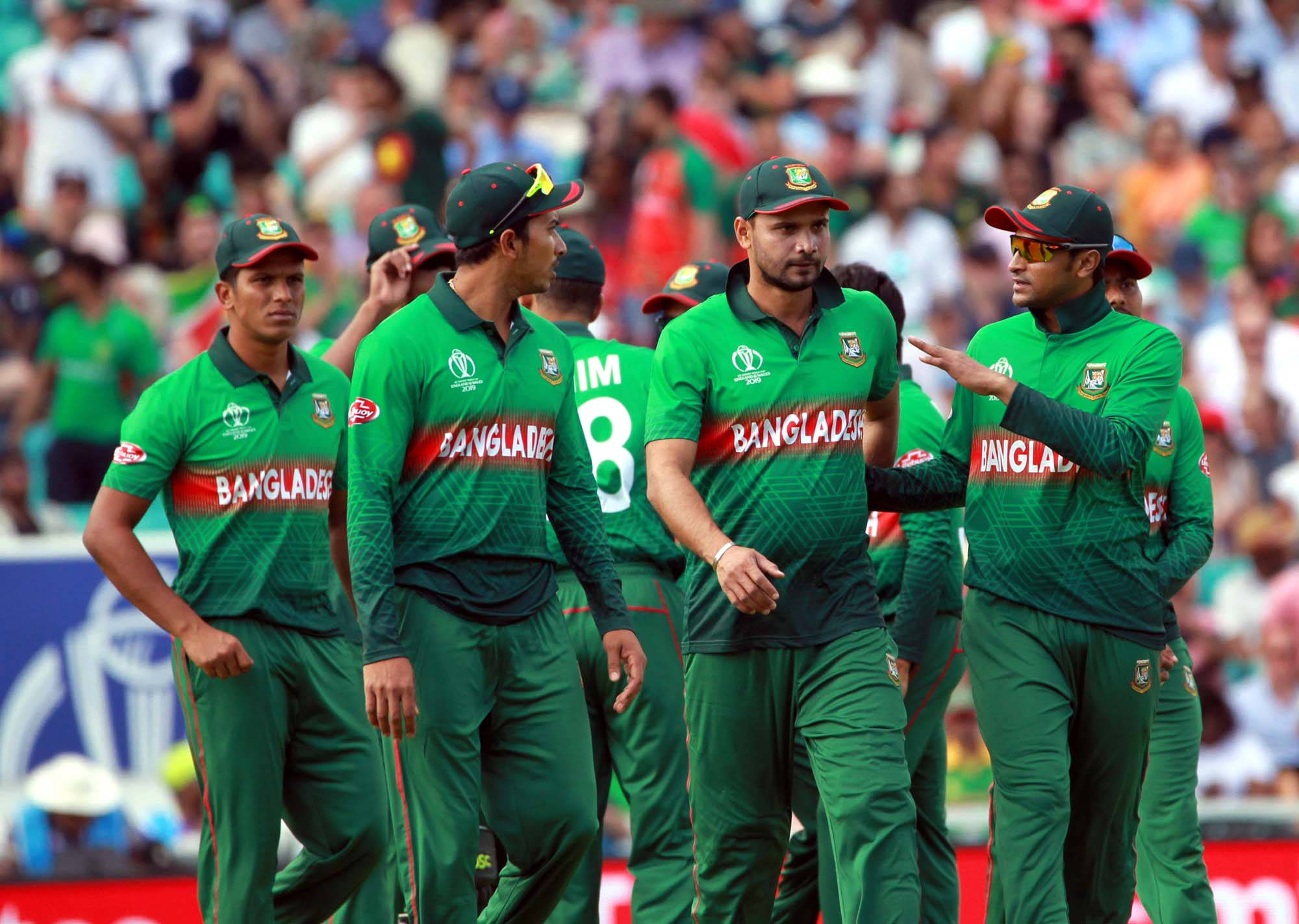Bangladesh Cricket 桌布 壁紙 — 孟加拉國板球隊球員身穿綠色制服，在陽光明媚的場地上聚在一起，背景是模糊的人群。