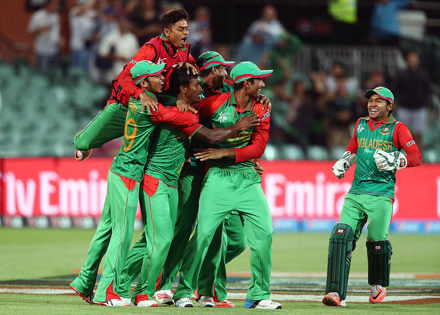 Bangladesh Cricket 桌布 壁紙 — 孟加拉國板球隊球員身穿綠色和紅色制服，在明亮的燈光下於體育場場地上以喜悅的擁抱慶祝勝利。